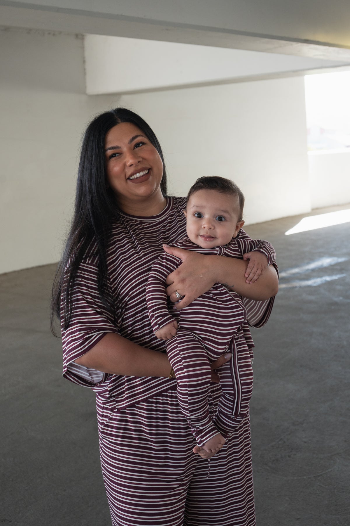 A smiling woman holds a baby, both dressed in matching forever french baby Bamboo Zip Pajamas in Maroon Stripe, made from breathable fabric, standing in a brightly lit indoor space.