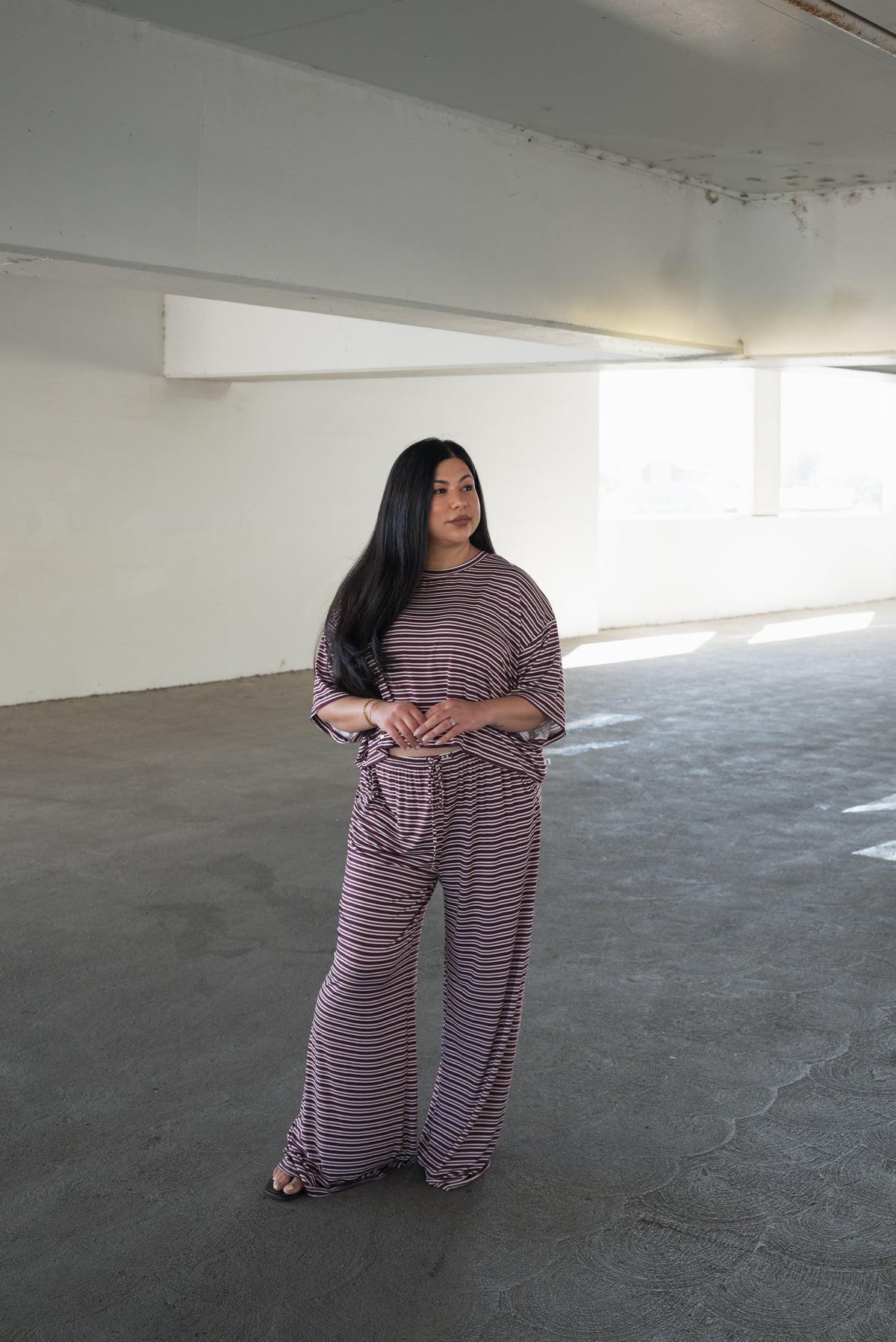 A woman with long dark hair stands in a bright, empty room, wearing the forever french baby Women’s Bamboo Crop Tee Lounge Set in Maroon Stripe. The breathable loungewear features a loose-fitting striped bamboo crop tee and matching wide-leg pant. She looks to the side with her hands loosely clasped in front.