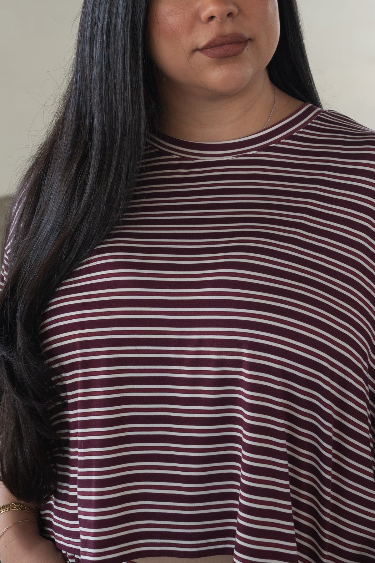 A woman with long dark hair wears the forever french baby Women’s Bamboo Crop Tee Lounge Set in Maroon Stripe. Only her face from the nose down and upper torso are visible. She has a neutral expression and accessorizes with a bracelet and a delicate necklace.