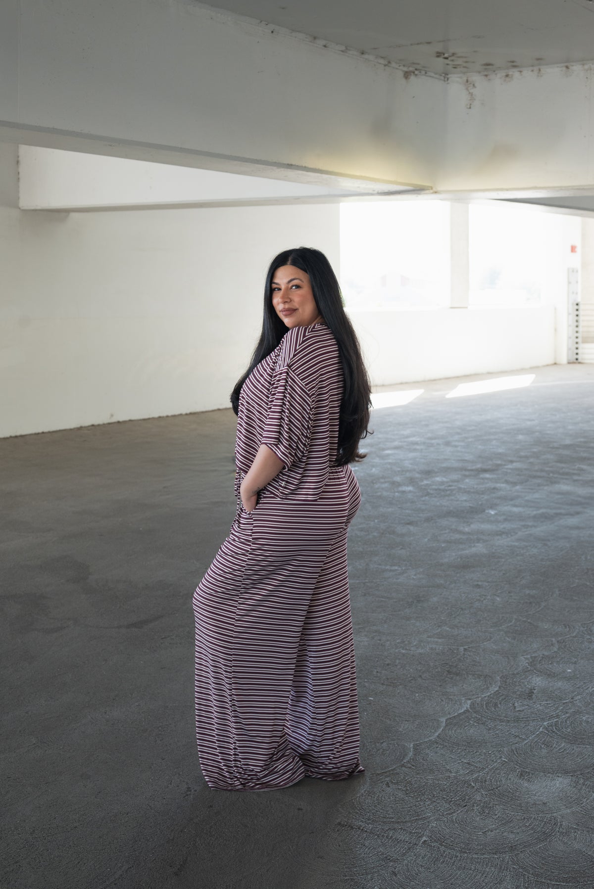 A woman with long dark hair stands in a bright, empty concrete space, looking over her shoulder at the camera while wearing the forever french baby Women’s Bamboo Crop Tee Lounge Set in Maroon Stripe—featuring a loose-fitting bamboo crop tee paired with wide-leg pants.