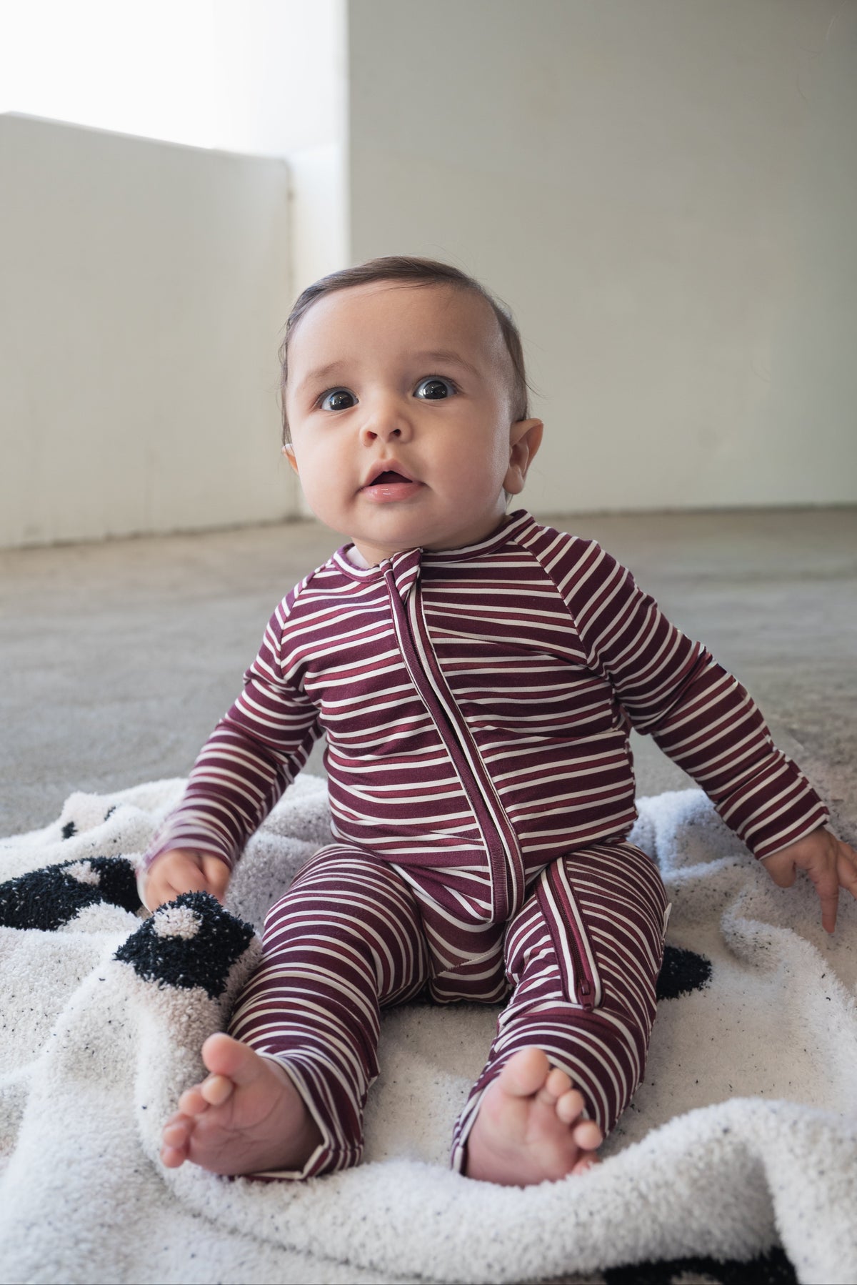 Dressed in the Bamboo Zip Pajamas | Maroon Stripe from forever french baby, a baby sits on a patterned blanket on the floor, gazing up with wide eyes.
