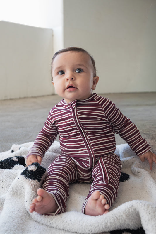 Dressed in the Bamboo Zip Pajamas | Maroon Stripe from forever french baby, a baby sits on a patterned blanket on the floor, gazing up with wide eyes.