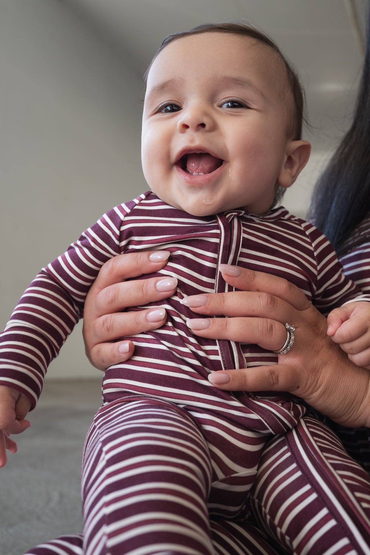 A smiling baby in the forever french baby Bamboo Zip Pajamas in Maroon Stripe is held by an adult whose hands, adorned with a ring, are visible. The cozy and breathable sleepwear is perfect for keeping your little one comfortable and happy.