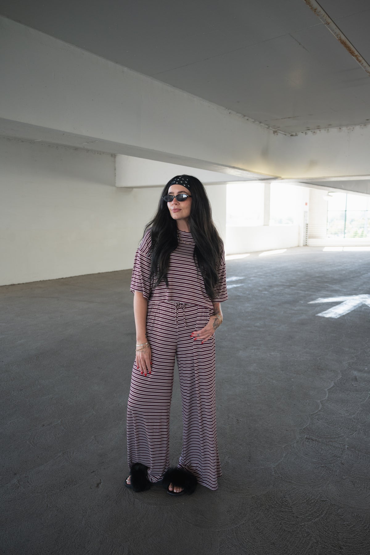 A woman in the forever french baby Women’s Bamboo Crop Tee Lounge Set in Maroon Stripe, complemented by a black headband and sunglasses, stands in a spacious, sunlit parking garage with concrete floors and white walls.