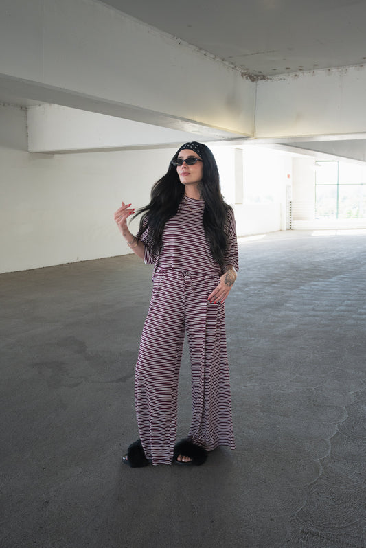 A woman in sunglasses and the forever french baby Women’s Bamboo Crop Tee Lounge Set in Maroon Stripe stands in a sunlit parking garage, looking to the side while holding her hair with one hand. Her relaxed bamboo crop tee and wide-leg pants offer breathable comfort and effortless style for warm days.