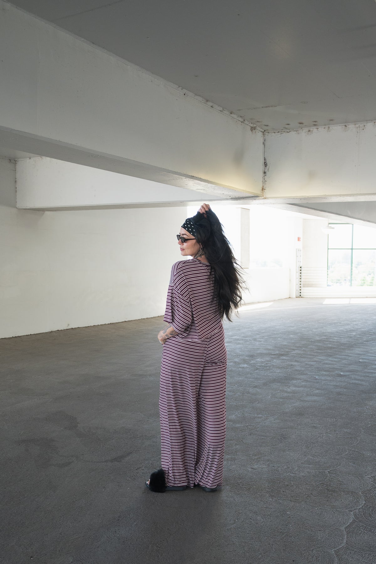 A woman in the forever french baby Women’s Bamboo Crop Tee Lounge Set in Maroon Stripe—featuring a wide-leg pant and matching crop tee—stands in a spacious, empty parking garage, facing away while looking back over her shoulder. She wears sunglasses and a black headband, holding up her long dark hair, effortlessly showcasing the comfort and breathability of this stylish loungewear set.