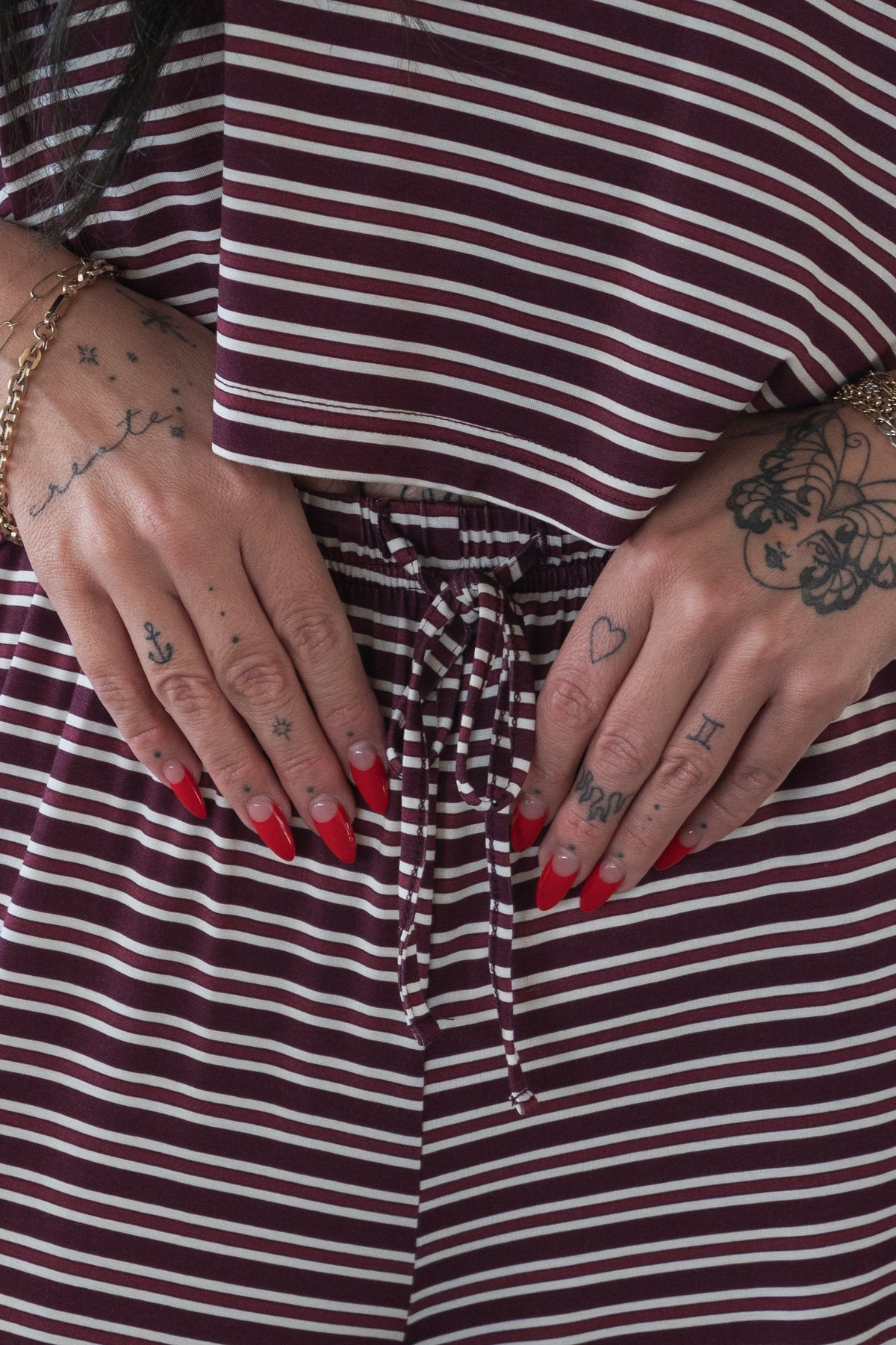 A person with red nails and hand tattoos rests their hands on the forever french baby Women’s Bamboo Crop Tee Lounge Set in Maroon Stripe, featuring wide-leg pants with a drawstring waist.