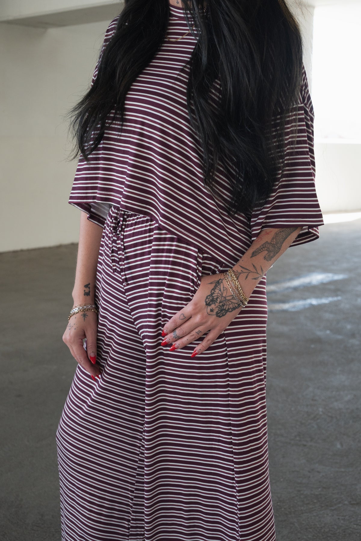 A person with long black hair, displaying tattooed arms and red painted nails, stands in a bright indoor space wearing the forever french baby Women’s Bamboo Crop Tee Lounge Set in Maroon Stripe—a comfortable and breathable loungewear ensemble.