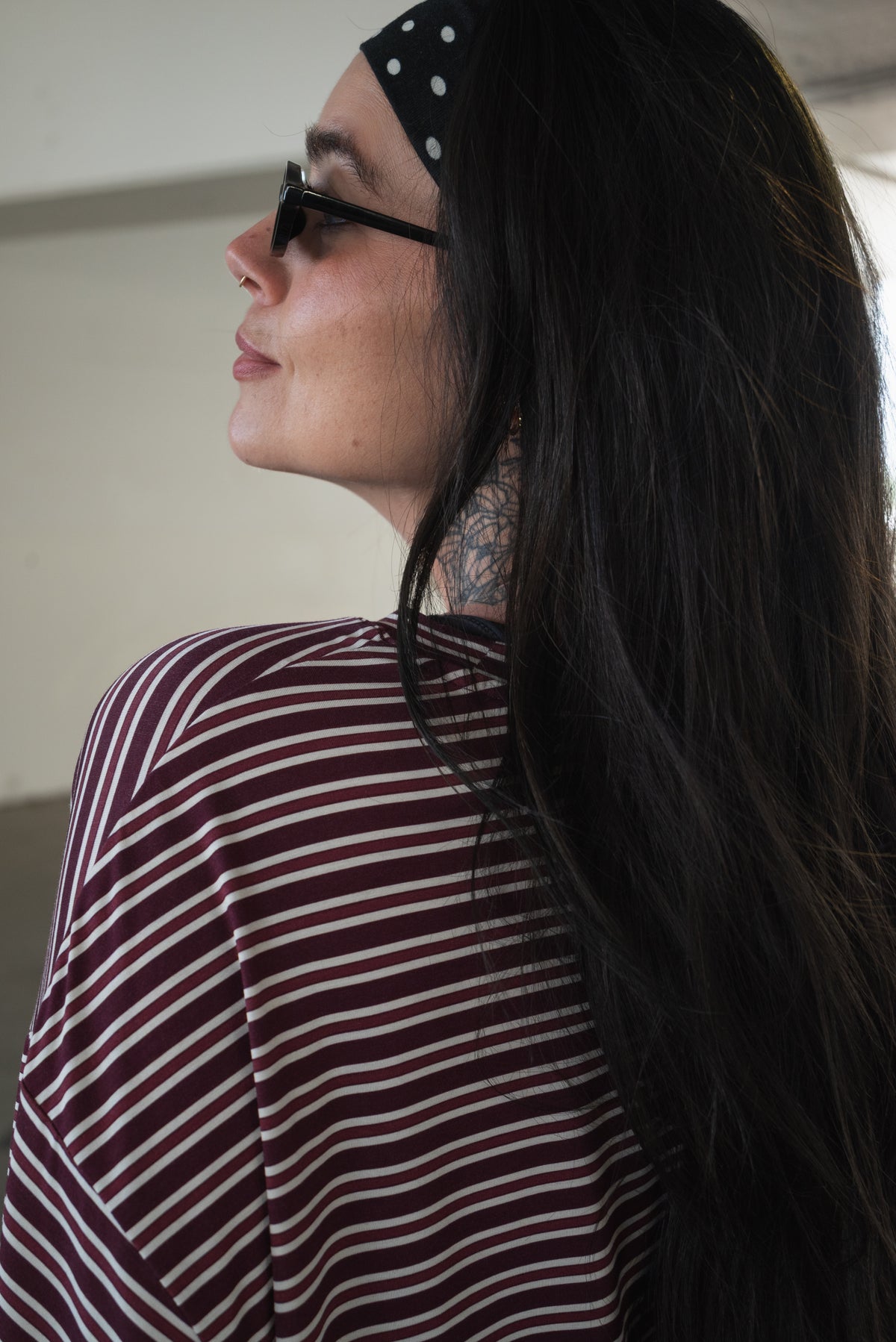 A person with long dark hair, wearing the forever french baby Women’s Bamboo Crop Tee Lounge Set in Maroon Stripe, paired with a black and white polka dot headband and sunglasses, is looking to the side in a softly lit indoor setting.