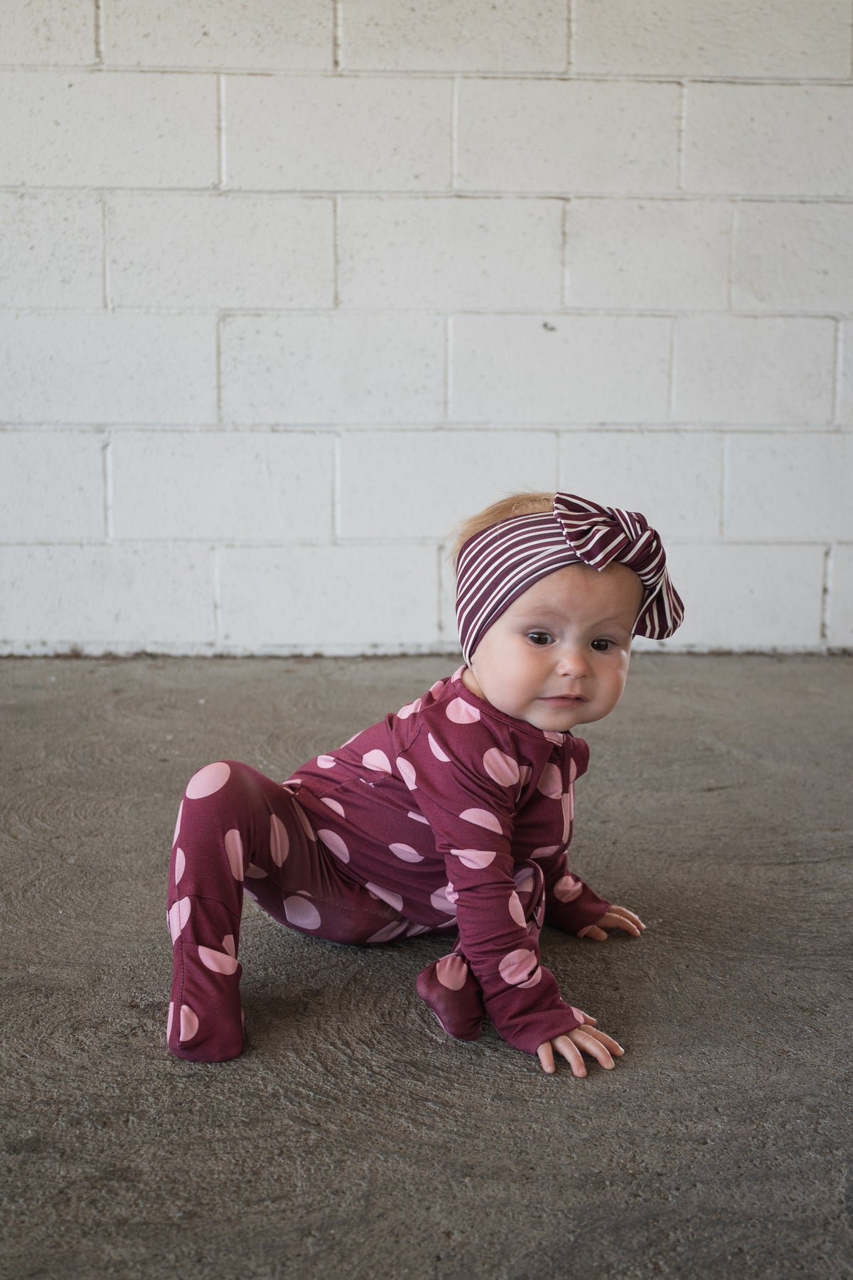 A baby wearing a maroon outfit with light pink spots and the forever french baby Bamboo Head Wrap in Maroon Stripe crawls on a concrete floor in front of a white brick wall.