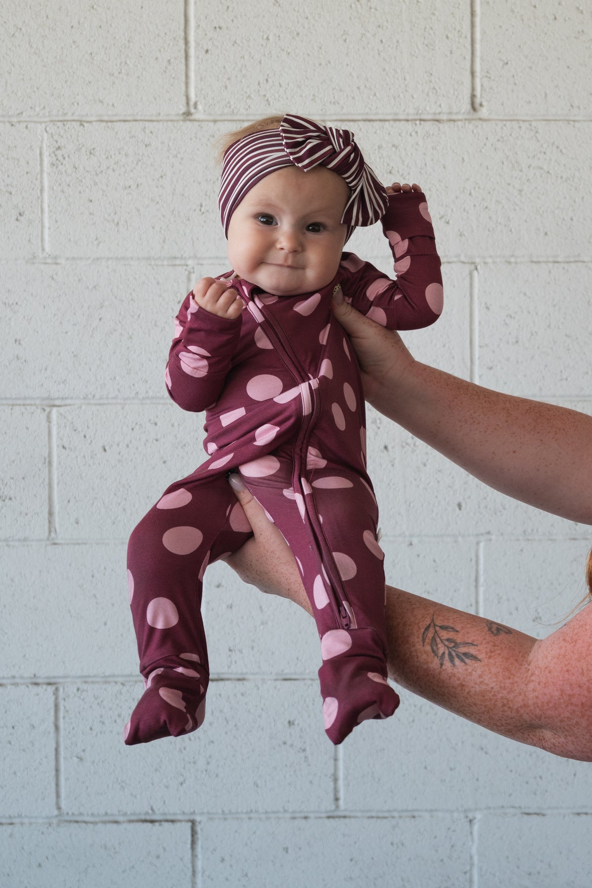 A baby wearing forever french baby Bamboo Zip Pajamas in Heather Rose Dot and a striped headband—made from hypoallergenic materials—is held up by an adult’s arm against a white brick wall.