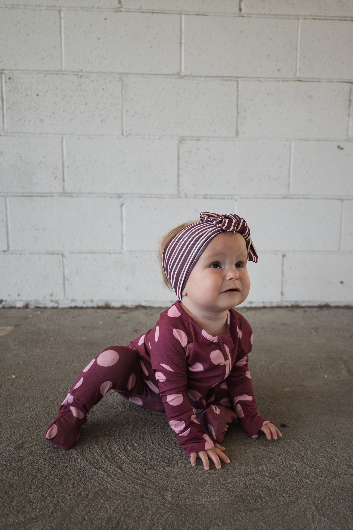 A baby dressed in forever french baby’s Bamboo Zip Pajamas in Heather Rose Dot, paired with a striped headband, crawls on a concrete floor before a white brick wall—showcasing the comfort and breathability of these stylish pajamas, perfect for little explorers.