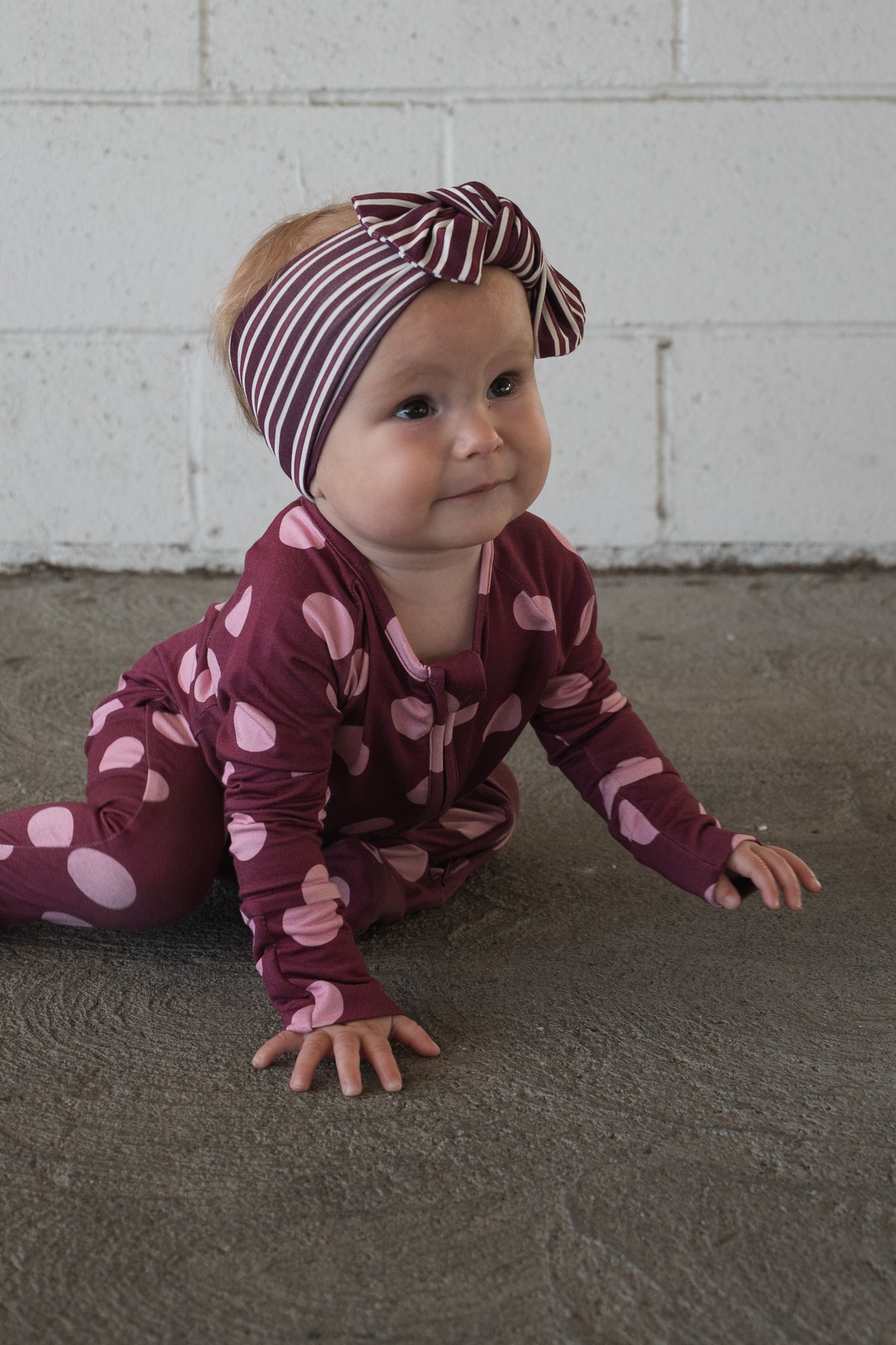 Dressed in forever french baby’s Bamboo Zip Pajamas in Heather Rose Dot, featuring a purple design with pink polka dots and a matching striped headband, a baby crawls on a gray concrete floor in front of a white brick wall.
