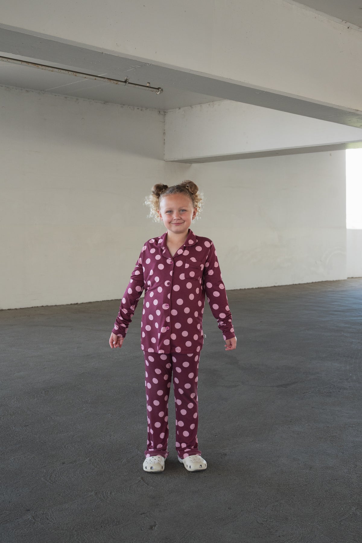 A young child with curly hair stands in an empty concrete room, smiling at the camera while wearing the forever french baby Girl’s Bamboo Button Up Lounge Set in Heather Rose Dot, featuring large white polka dots and paired with cozy white slippers. The soft, hypo-allergenic bamboo fabric brings both comfort and style to her cheerful look.