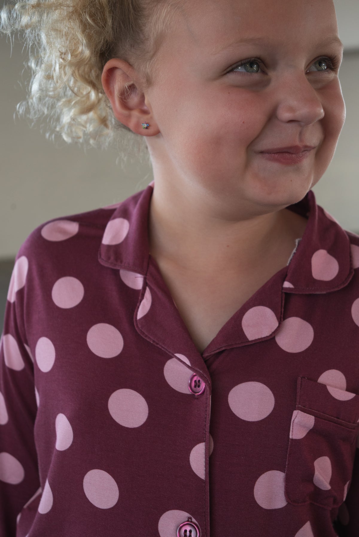 A young child with curly blond hair smiles while wearing the forever french baby Girl’s Bamboo Button Up Lounge Set in Heather Rose Dot, enjoying the comfort of its soft, hypo-allergenic bamboo fabric.