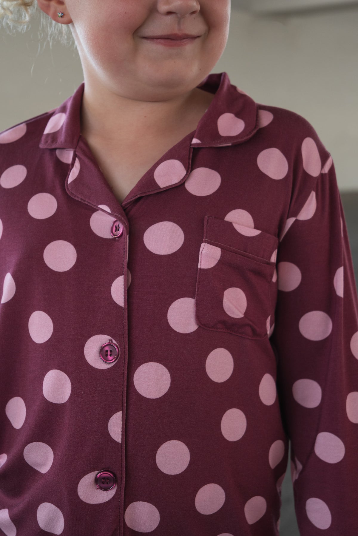 A child wearing forever french baby’s Girl’s Bamboo Button Up Lounge Set in Heather Rose Dot, featuring a maroon base with large pink polka dots, shows only the upper body and a smiling mouth. The soft, hypo-allergenic bamboo fabric ensures both comfort and style.