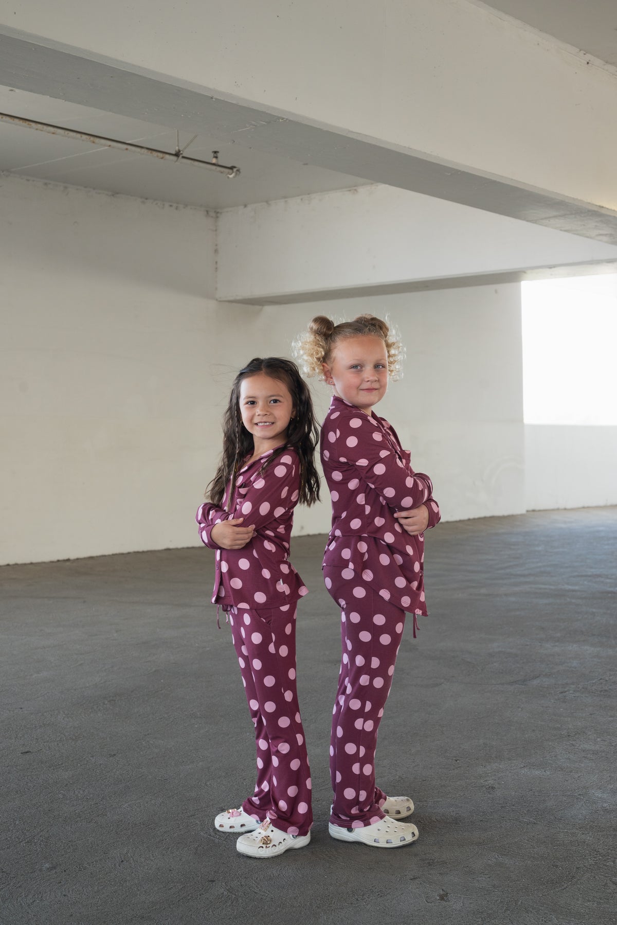 Two young girls stand back-to-back, smiling and wearing matching forever french baby Girl’s Bamboo Button Up Lounge Sets in Heather Rose Dot with white shoes, in a large, empty, brightly-lit room. Their hypo-allergenic bamboo outfits look comfortable and perfect for playful moments together.