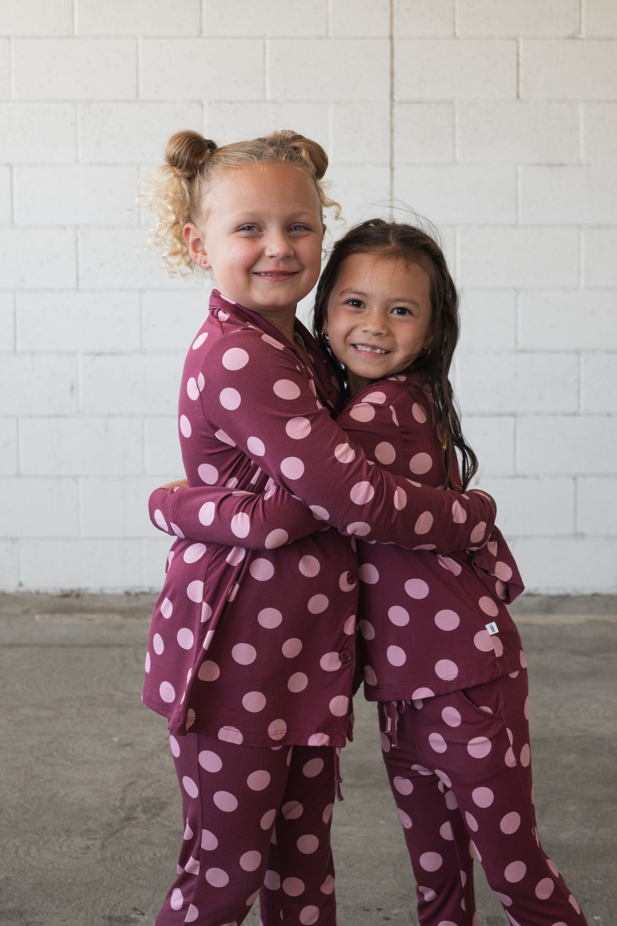 Two young children wearing matching forever french baby Girl’s Bamboo Button Up Lounge Sets in Heather Rose Dot hug each other and smile, standing in front of a white brick wall. Their soft, hypoallergenic bamboo outfits keep them cozy and comfortable as they share this sweet moment.