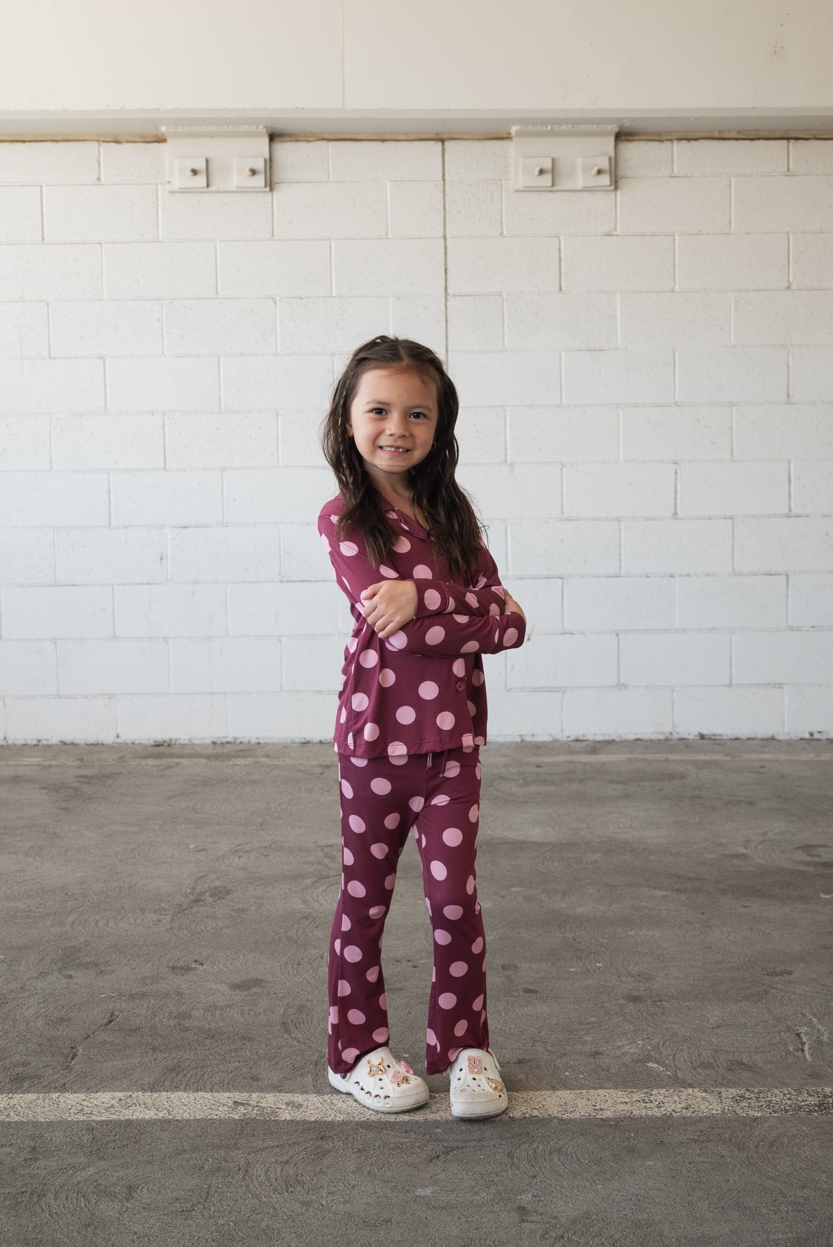 A young girl with long dark hair stands confidently with arms crossed, wearing the forever french baby Girl’s Bamboo Button Up Lounge Set in Heather Rose Dot—a stylish maroon set adorned with large pink polka dots—paired with white shoes, against a white brick wall in a parking area. Made from hypo-allergenic bamboo fabric, this lounge set is perfect for sensitive skin.
