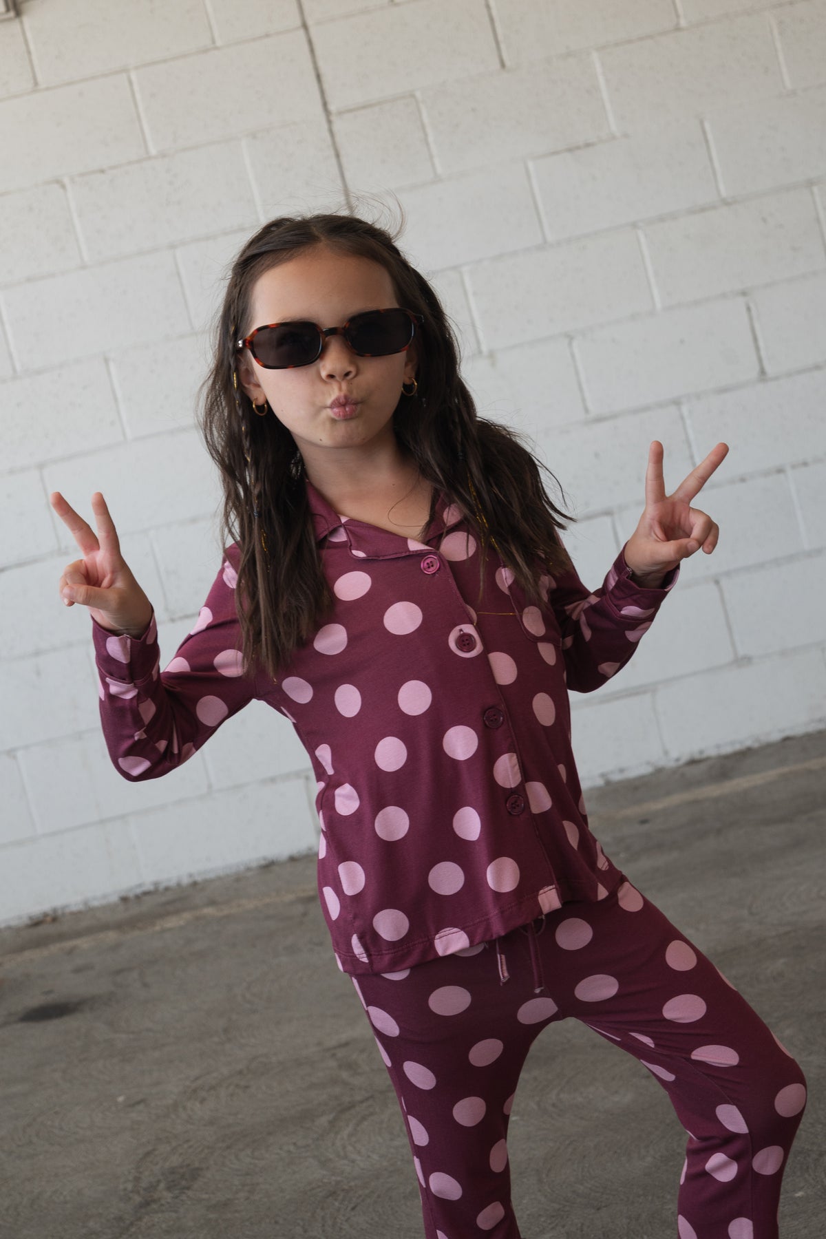 A young girl wearing sunglasses and the forever french baby Girl’s Bamboo Button Up Lounge Set in Heather Rose Dot poses with both hands making peace signs in front of a white brick wall. This hypo-allergenic bamboo set ensures comfort and style for any playful moment.