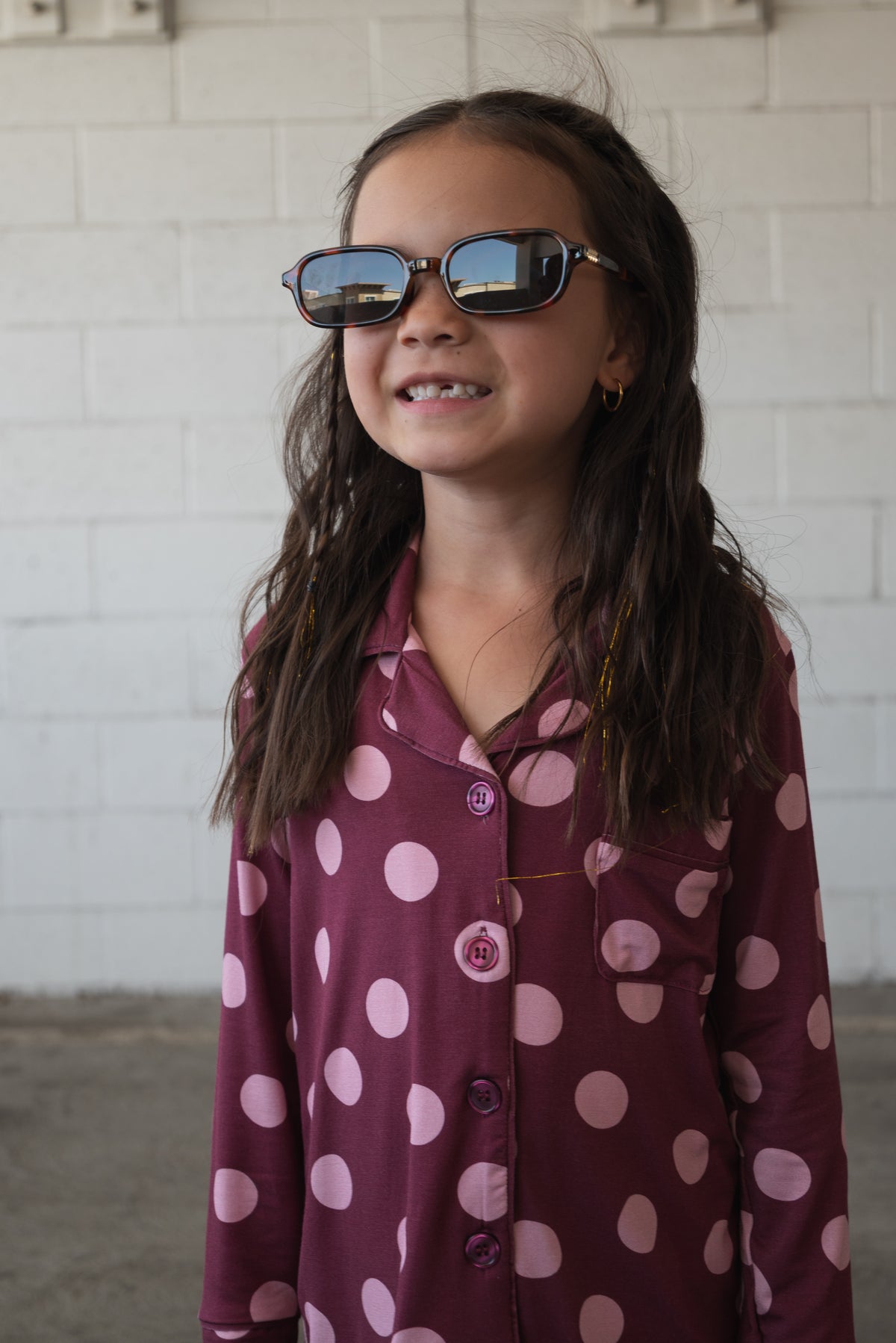 A young girl with long brown hair, wearing sunglasses and the Girl’s Bamboo Button Up Lounge Set in Heather Rose Dot from forever french baby, smiles while standing in front of a white brick wall. The hypo-allergenic bamboo fabric ensures she stays comfortable and stylish during all her everyday adventures.