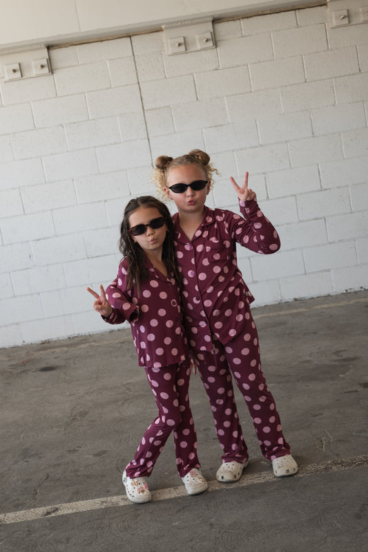 Two children wearing matching forever french baby Girl’s Bamboo Button Up Lounge Set in Heather Rose Dot, paired with sunglasses and white clogs, pose together against a white brick wall, each flashing a peace sign. This adorable lounge set is crafted from hypo-allergenic bamboo fabric, ensuring ultimate comfort.