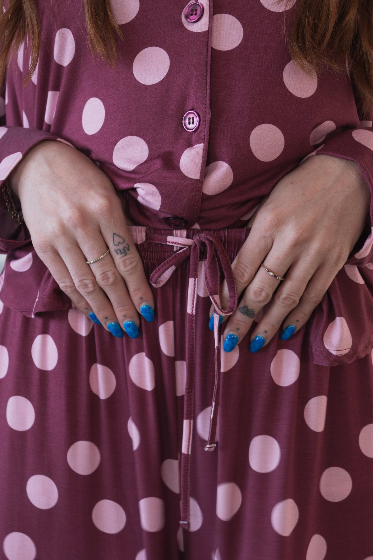 A person with blue-painted nails, rings, and small finger tattoos rests their hands on their waist while wearing the forever french baby Women’s Bamboo Button Up Lounge Set in Heather Rose Dot. This purple set features large pink polka dots and a tie at the waist.