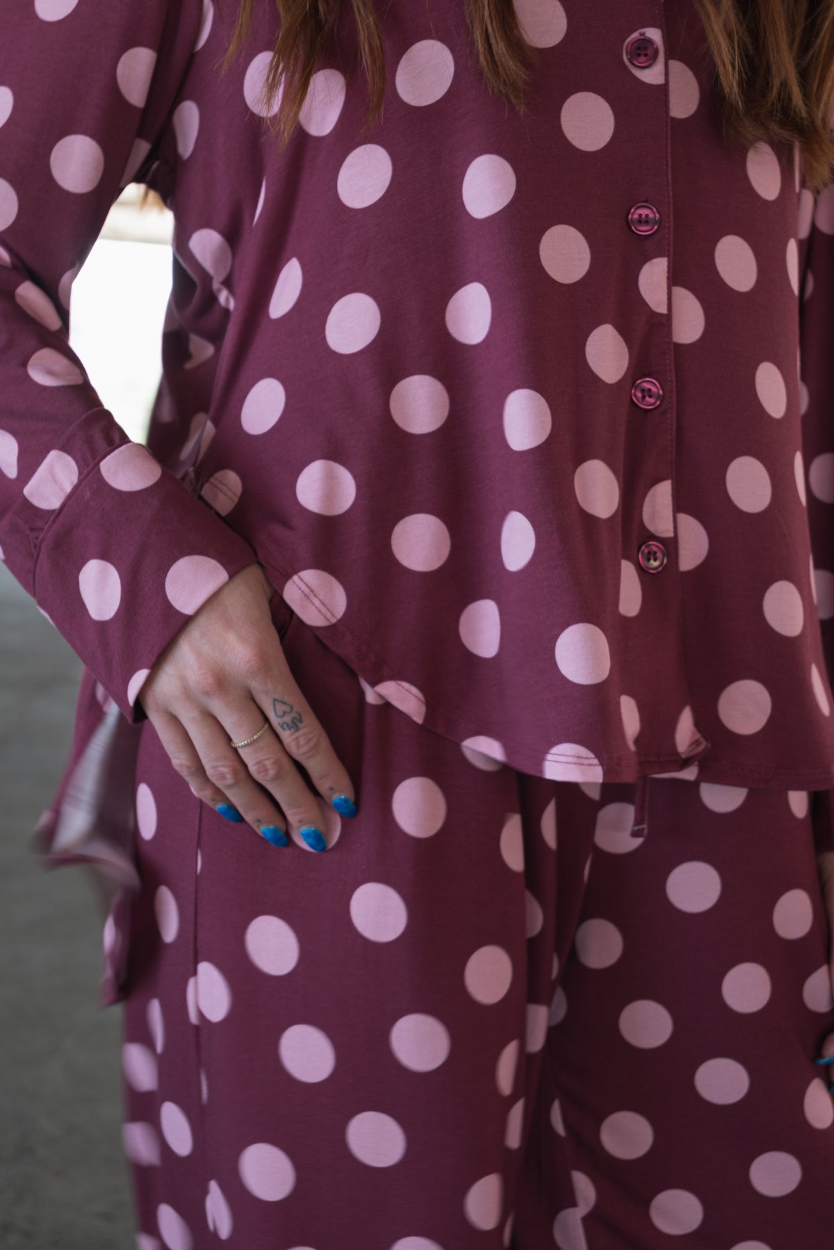 Clad in the forever french baby Women’s Bamboo Button Up Lounge Set in Heather Rose Dot—featuring a matching maroon top and wide leg pants decorated with large pink polka dots—a person lounges comfortably, their blue nail polished hand with a small tattoo resting on their thigh.