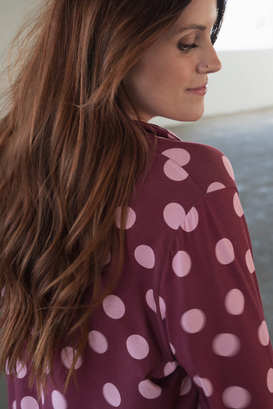 A woman with long brown hair, dressed in the forever french baby Women’s Bamboo Button Up Lounge Set in Heather Rose Dot—a maroon set featuring large pink polka dots and wide leg lounge pants—looks down with a soft smile in a gently lit indoor setting.