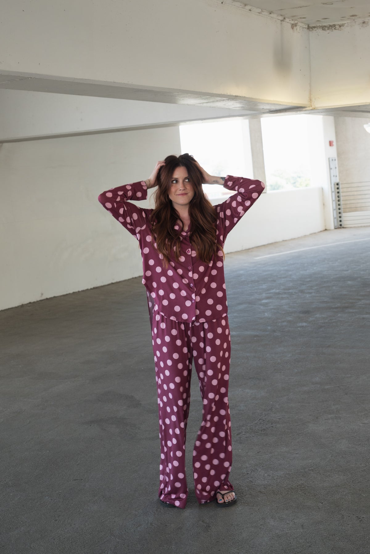 A woman stands on a concrete floor in a parking garage, wearing the forever french baby Women’s Bamboo Button Up Lounge Set in Heather Rose Dot—a purple set with white polka dots and wide leg lounge pants—paired with sandals, as she holds her hands to her head and gazes forward.