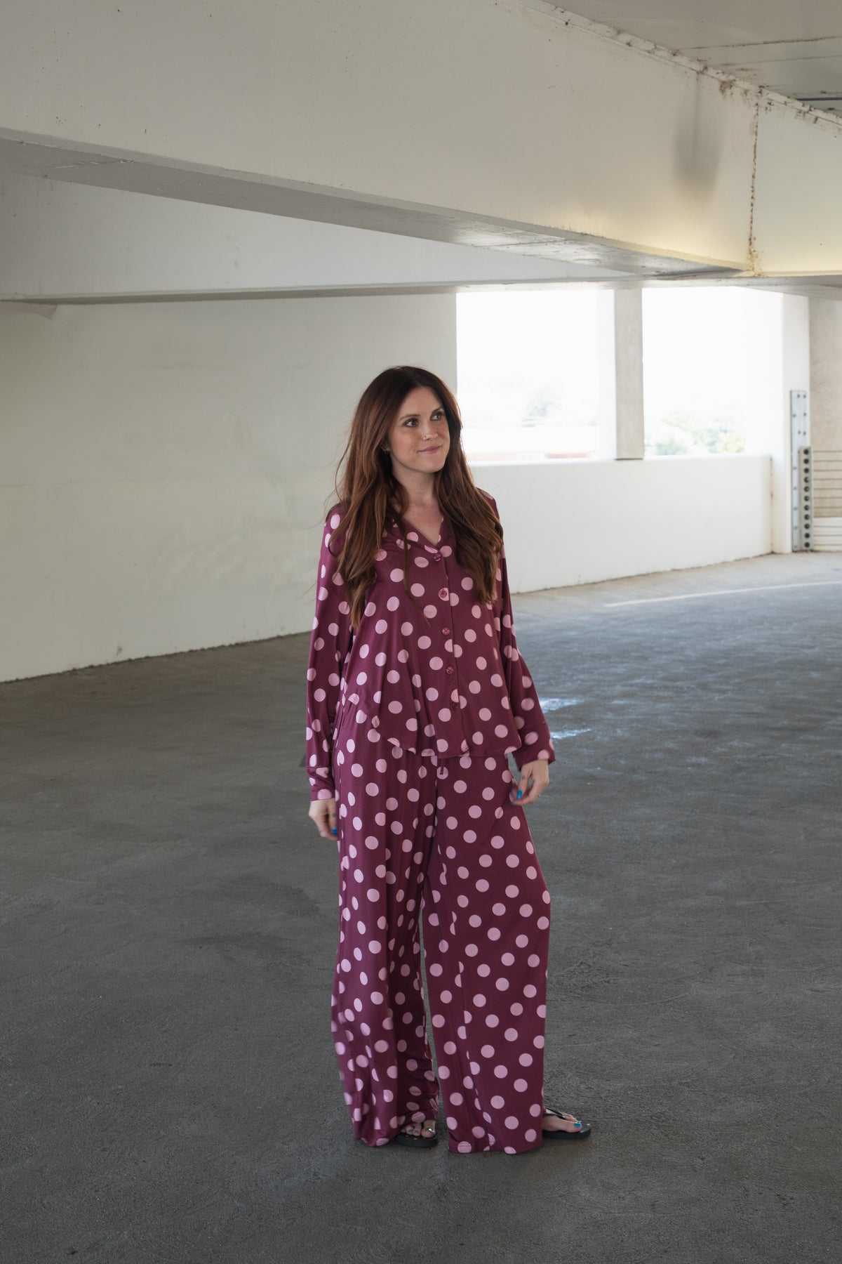 A woman stands in a parking garage wearing the forever french baby Women’s Bamboo Button Up Lounge Set in Heather Rose Dot. She has long brown hair and gazes slightly to the left with a calm expression, her wide leg lounge pants lending a relaxed and stylish vibe to her bamboo clothing ensemble.