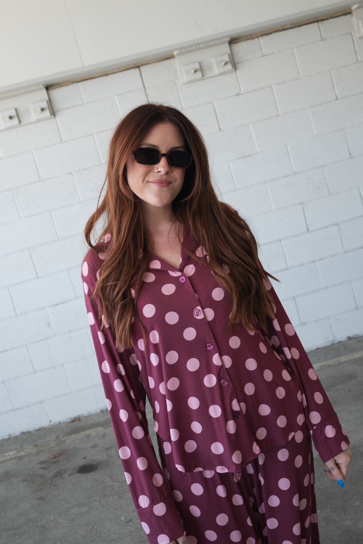 A woman with long brown hair, wearing black sunglasses and the Women’s Bamboo Button Up Lounge Set in Heather Rose Dot by forever french baby, featuring wide leg lounge pants, stands in front of a white brick wall, smiling slightly.