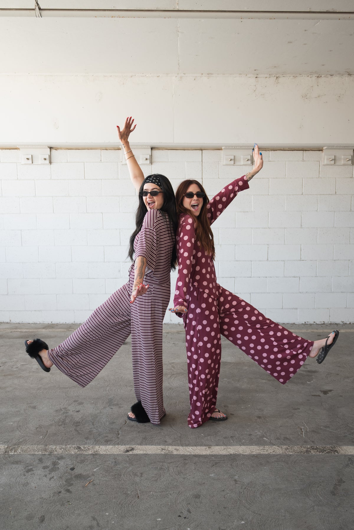 Two people in matching forever french baby Women’s Bamboo Button Up Lounge Sets in Heather Rose Dot pose back-to-back, each standing on one leg with arms raised and tongues out, exuding a cheerful, playful vibe against a white brick wall in a parking garage.