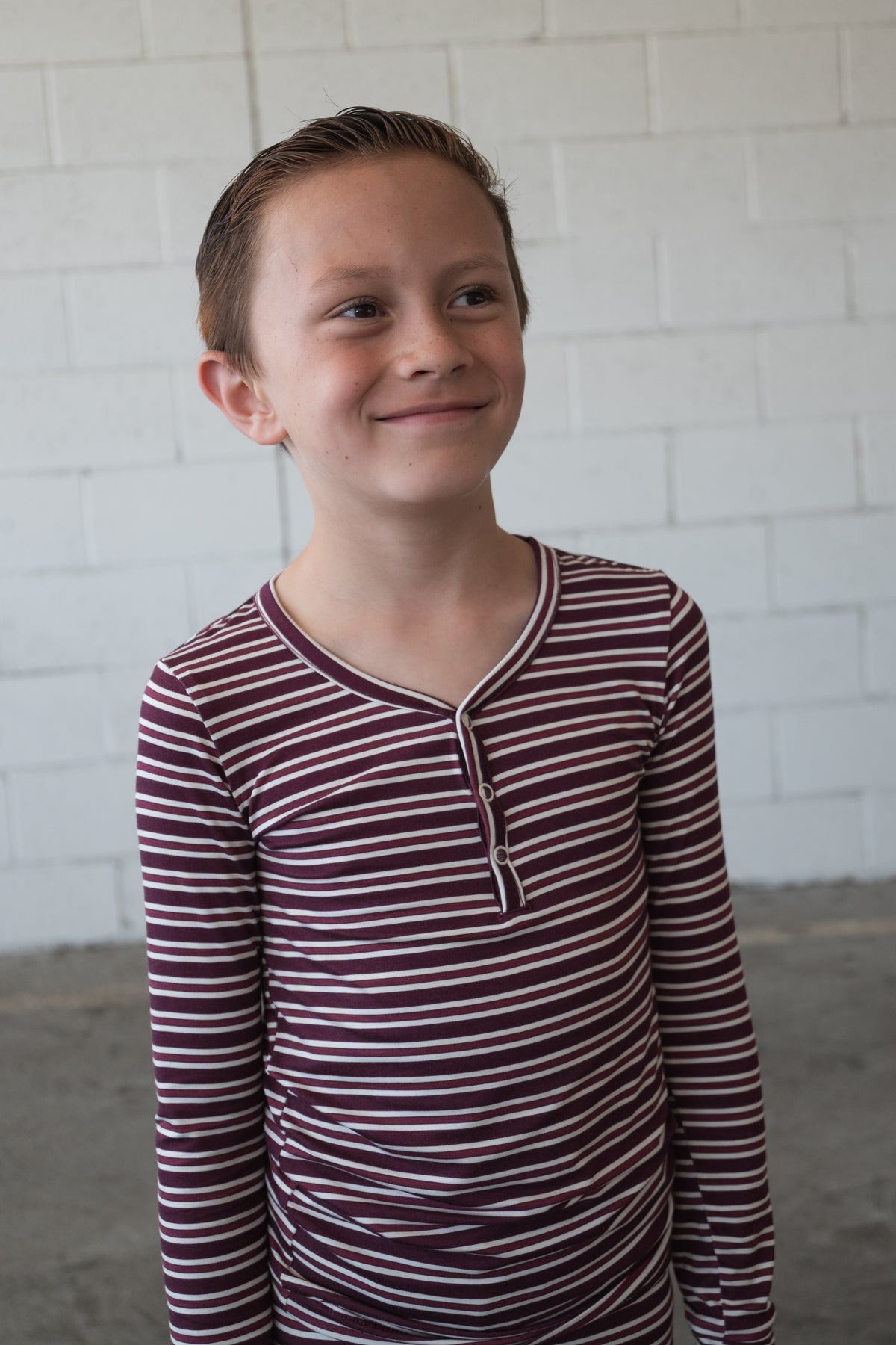 A young child with short brown hair is wearing the forever french baby Bamboo Two Piece Pajamas in Maroon Stripe, standing in front of a white brick wall and smiling slightly while looking to the side.