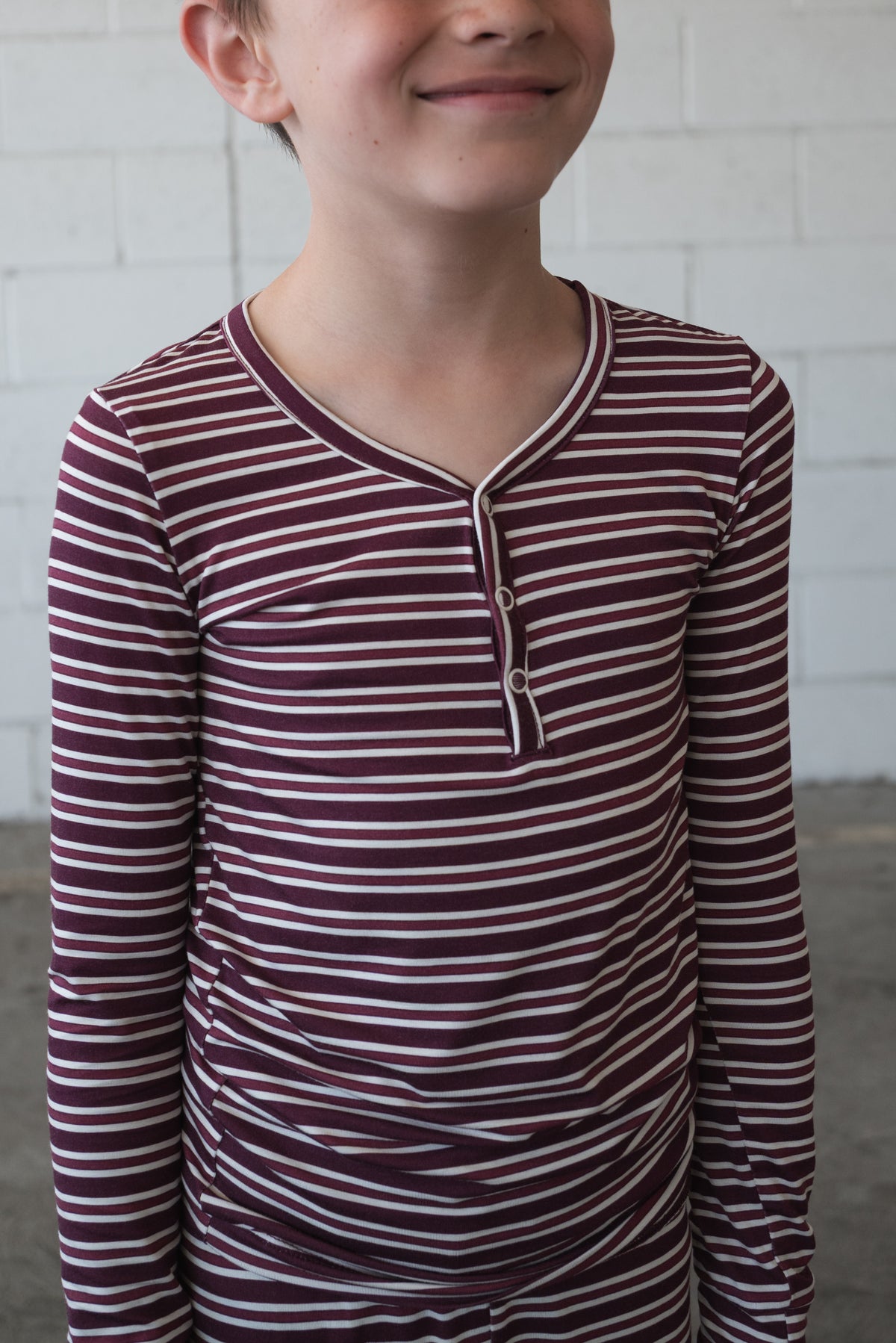 A child wearing the forever french baby Bamboo Two Piece Pajamas in Maroon Stripe smiles in front of a white brick wall, with only the lower part of their face visible.
