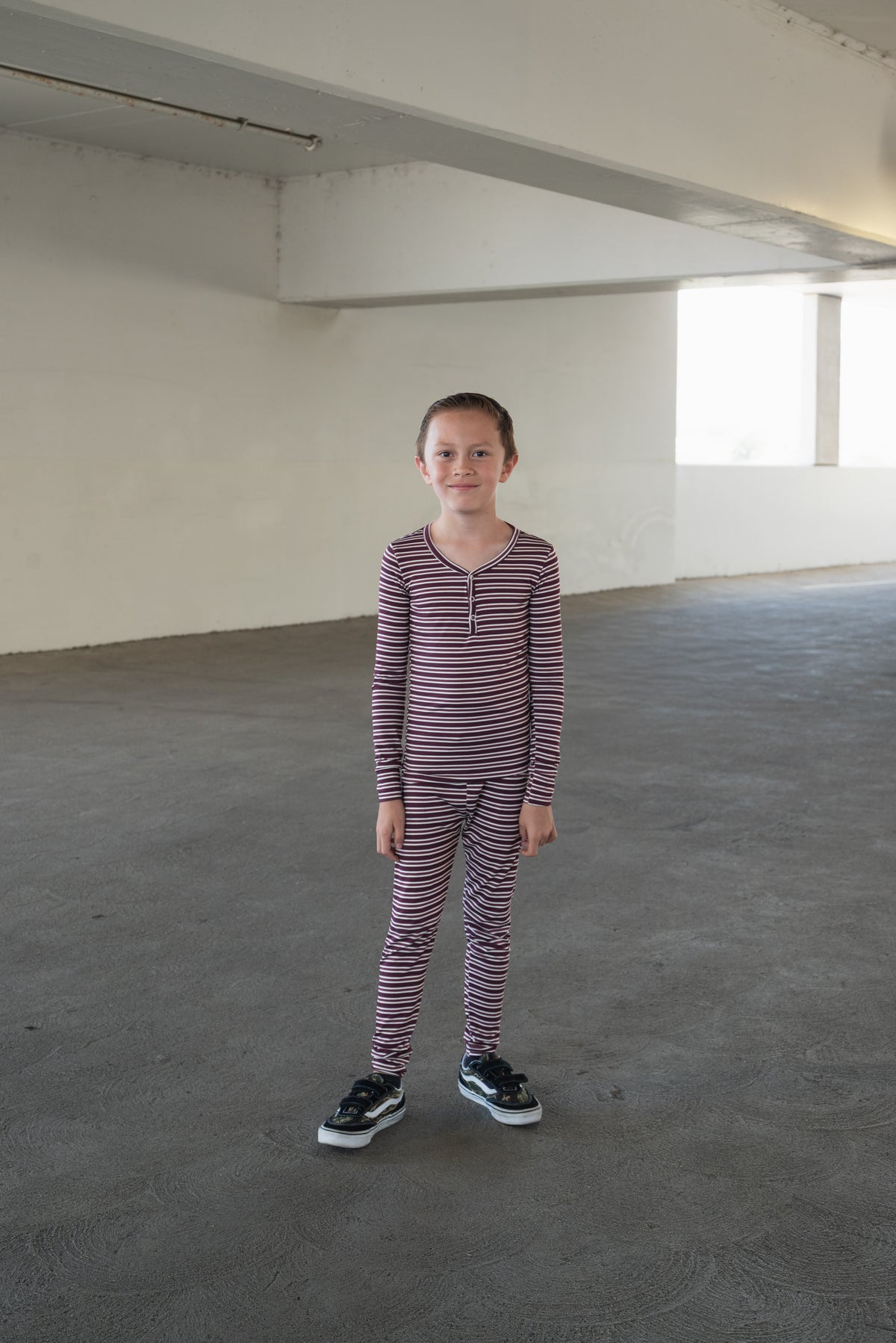 A child wearing the forever french baby Bamboo Two Piece Pajamas in Maroon Stripe stands in a spacious, empty concrete room with natural light streaming through a window, cozy and comfortable in breathable kids sleepwear perfect for restful nights.