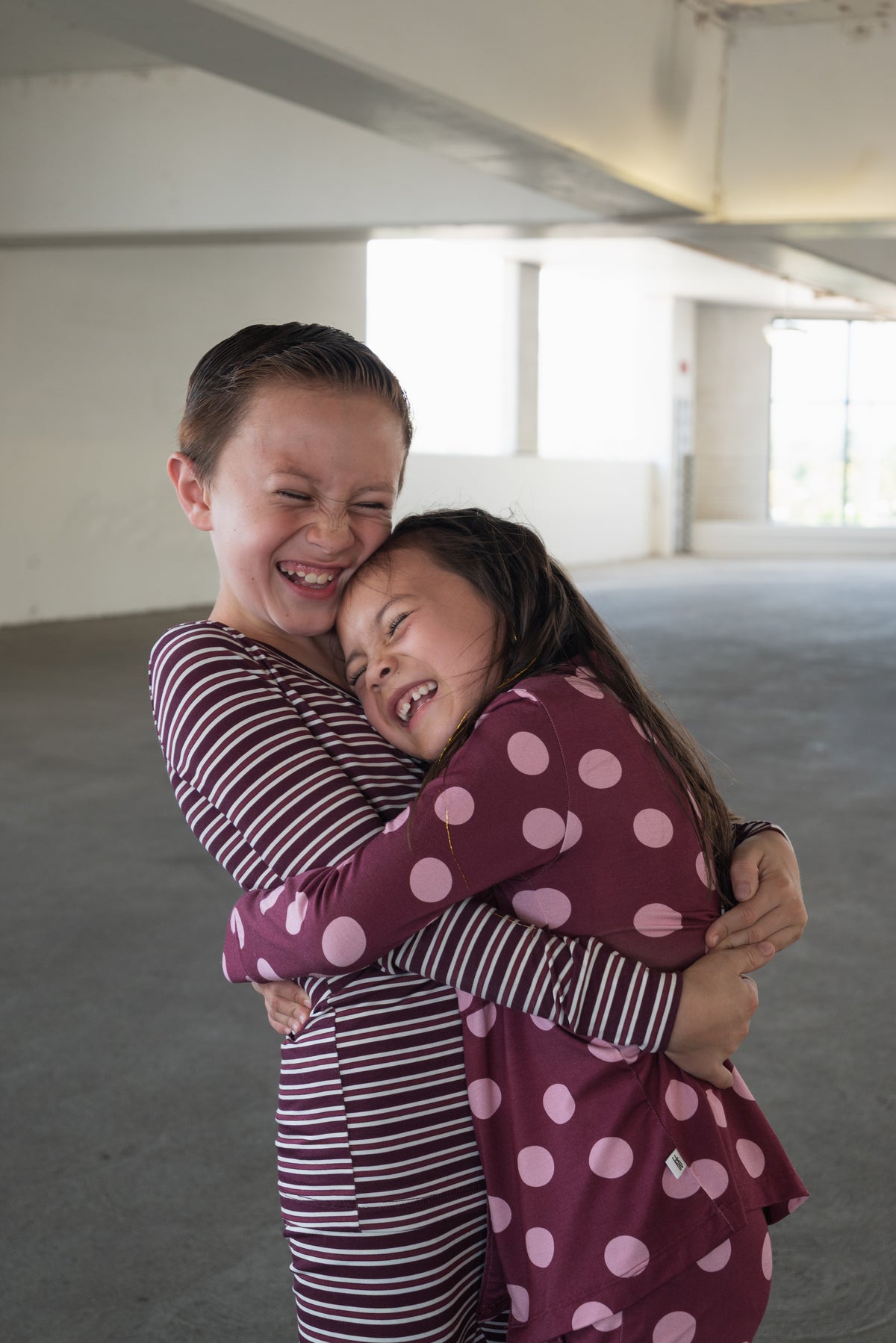 Wearing matching maroon outfits, two children embrace and smile brightly in a sunlit indoor space. The girl’s forever french baby Bamboo Button Up Lounge Set in Heather Rose Dot is not only charming but also hypo-allergenic, ensuring playtime is comfortable and carefree.