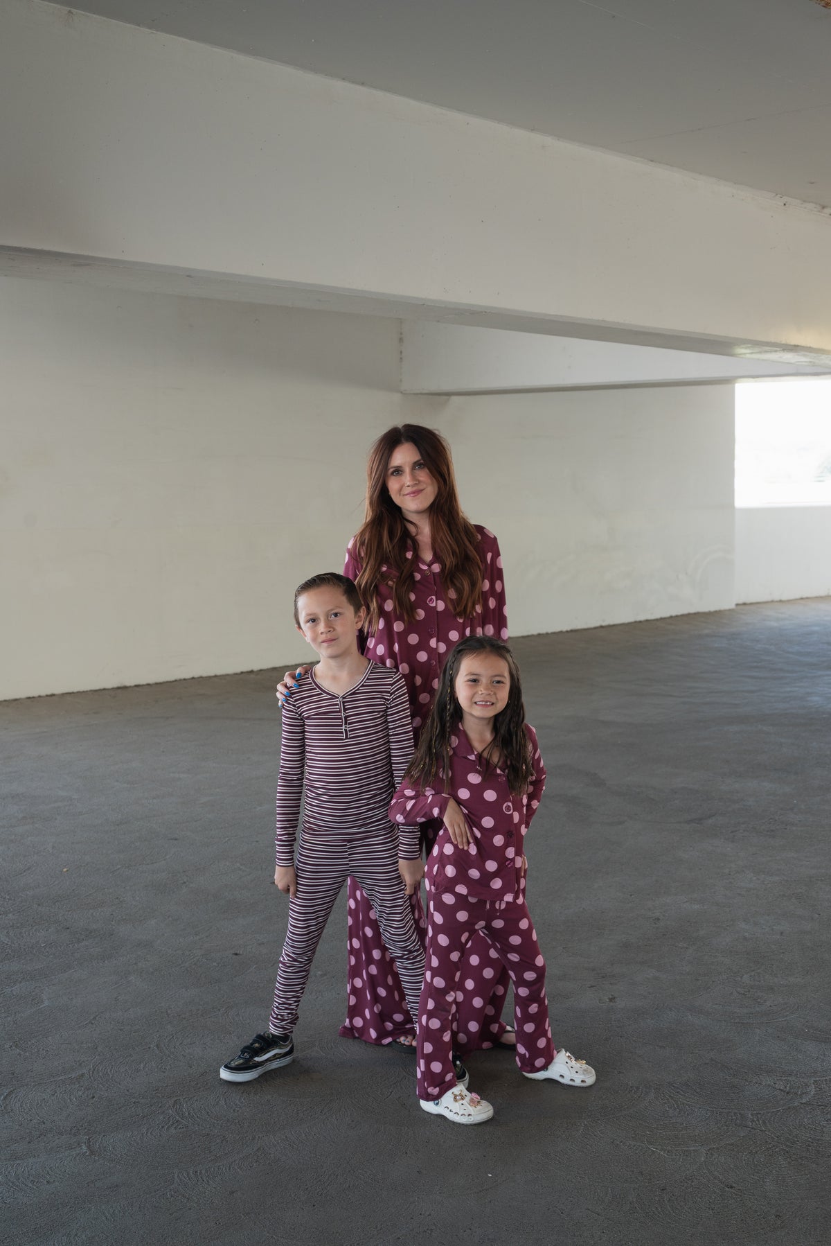 A woman wearing the forever french baby Women’s Bamboo Button Up Lounge Set in Heather Rose Dot stands behind two girls—one dressed in stripes and the other in polka dots—as they pose together under a concrete structure. Their playful style is enhanced by cozy bamboo clothing, making it perfect for relaxed days and casual gatherings.