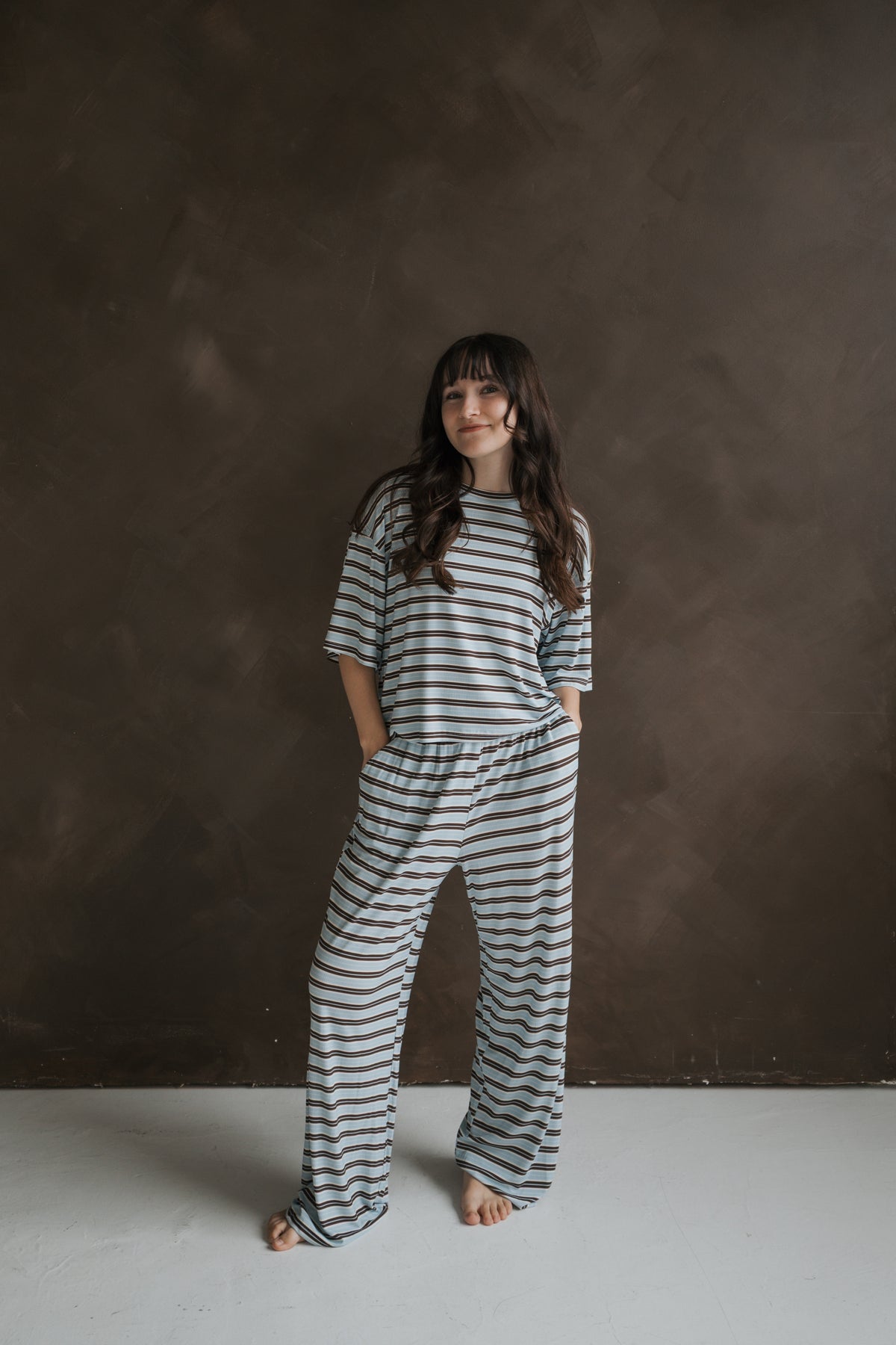 A woman wearing the forever french baby Women’s Boxy Tee Lounge Set | Dean, featuring a matching short-sleeved top and wide-leg trousers in light blue and brown horizontal stripes of breathable bamboo loungewear.