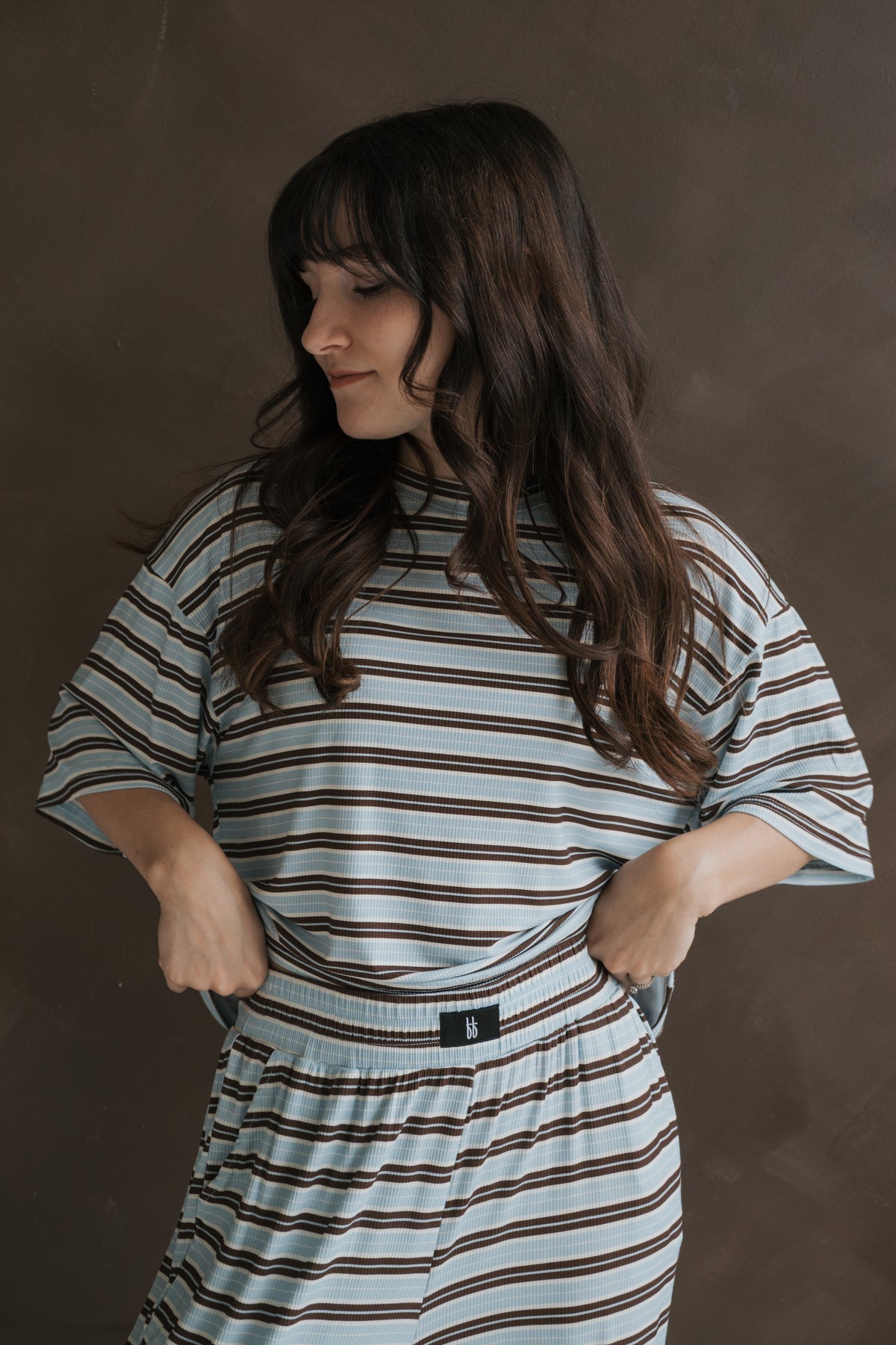 A woman wears a Women’s Boxy Tee Lounge Set | Dean by forever french baby, featuring breathable, bamboo-based loungewear with a boxy, short-sleeved top and matching lounge pants in a blue, white, and brown horizontal stripe pattern.