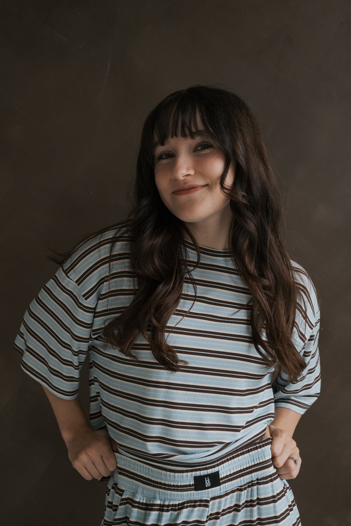 A woman wears a Women’s Boxy Tee Lounge Set | Dean, featuring a breathable, boxy short-sleeved top and elastic-waist shorts with blue and brown horizontal stripes.