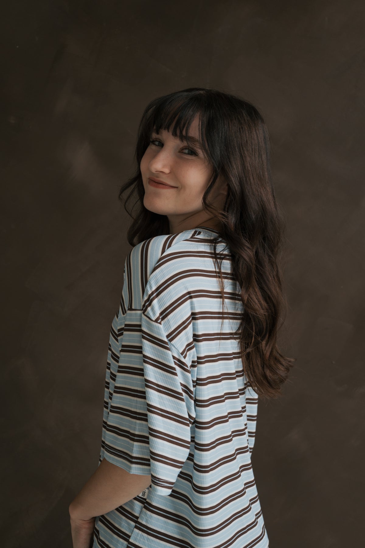 A woman wears the Women’s Boxy Tee Lounge Set | Dean, a boxy, short-sleeved shirt with brown and light blue horizontal stripes made of breathable bamboo clothing.