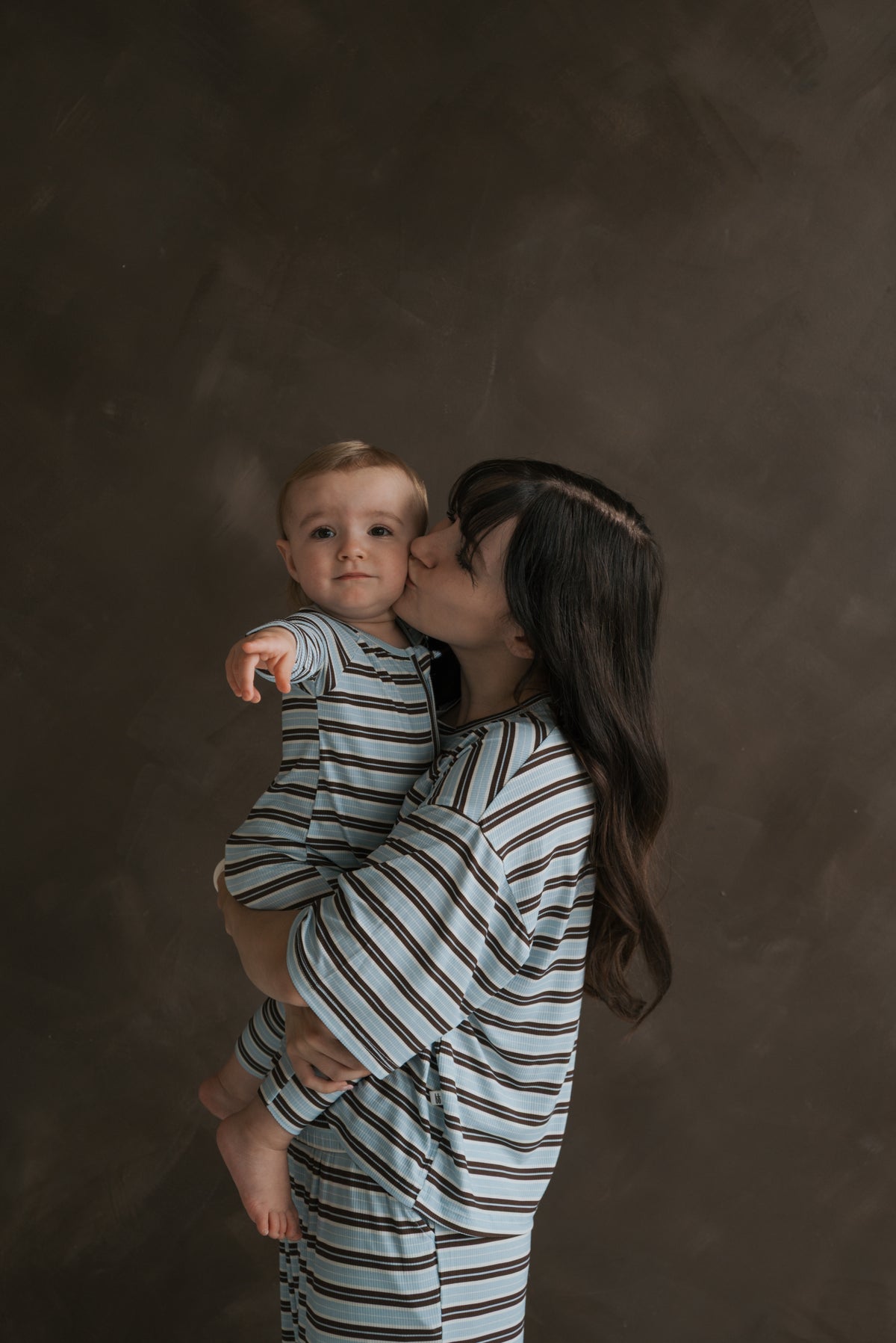 A woman in a Women’s Boxy Tee Lounge Set | Dean kisses her baby’s cheek as they both wear matching blue and brown striped bamboo loungewear.