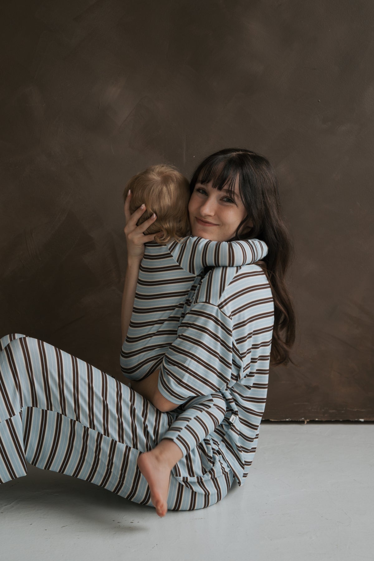 A woman sits on the floor holding a young child in her lap, both wearing matching blue and brown ribbed Bamboo Zip Pajamas | Dean by forever french baby.