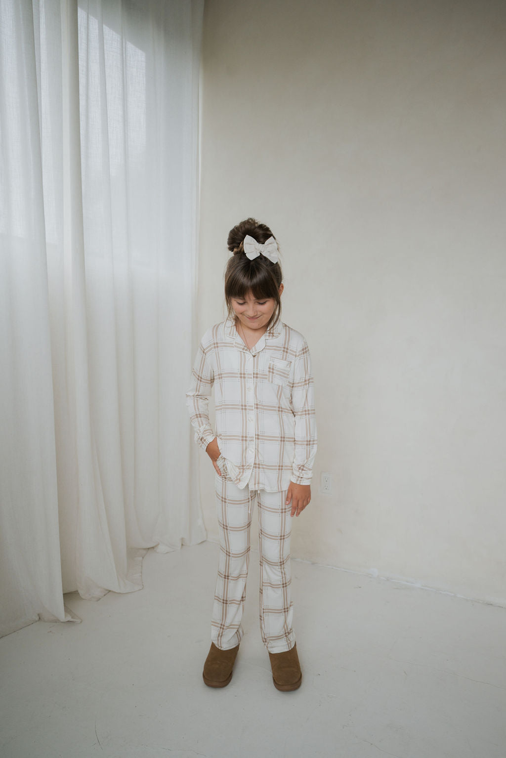 A girl wearing a Girl’s Bamboo Button Up Lounge Set | Ellis Plaid, featuring a long-sleeved, button-up top and pants in a light tan and white plaid pattern.