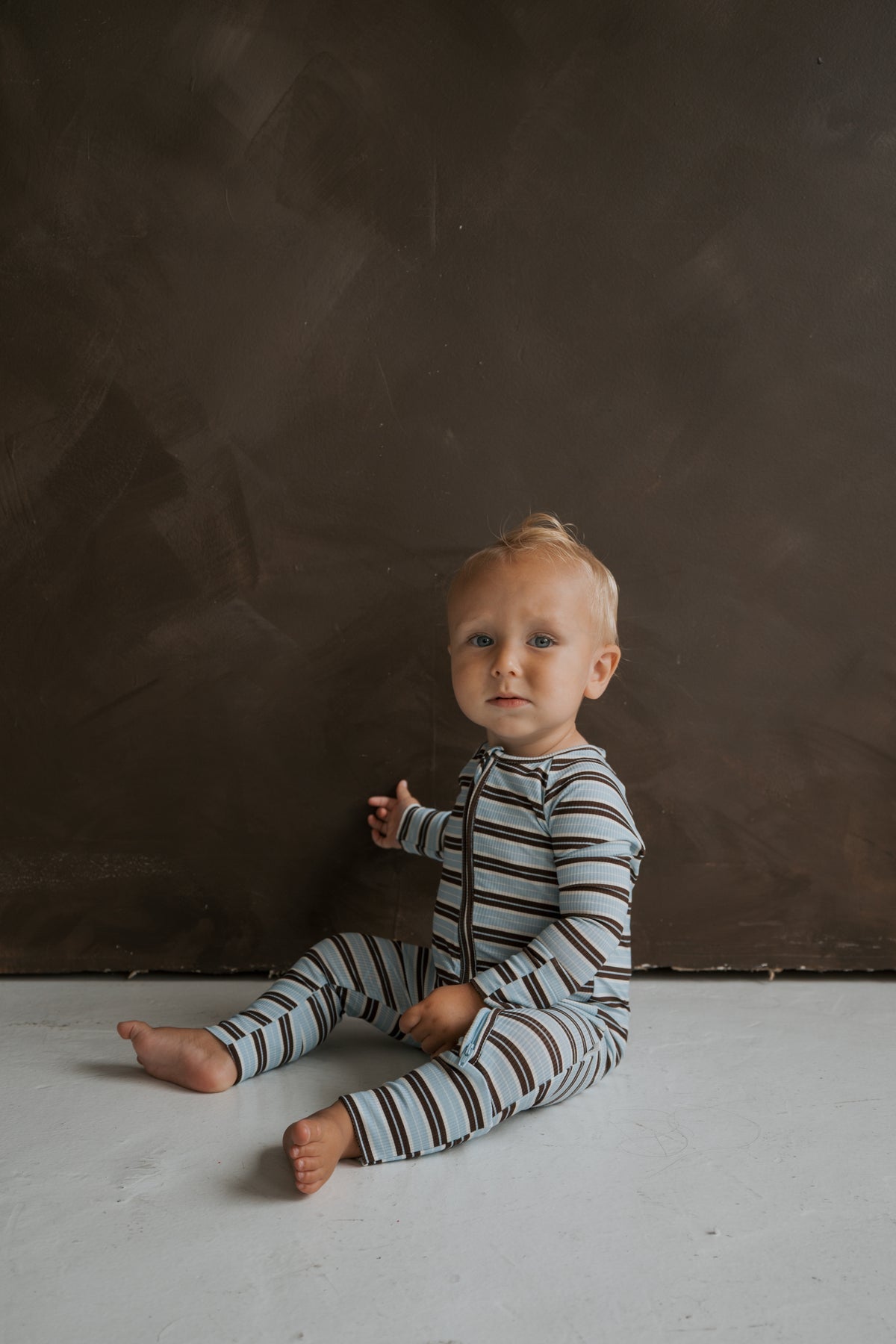 A baby wearing long-sleeved, footless ribbed Bamboo Zip Pajamas | Dean in blue and brown horizontal stripes, sitting on a white floor against a brown wall.