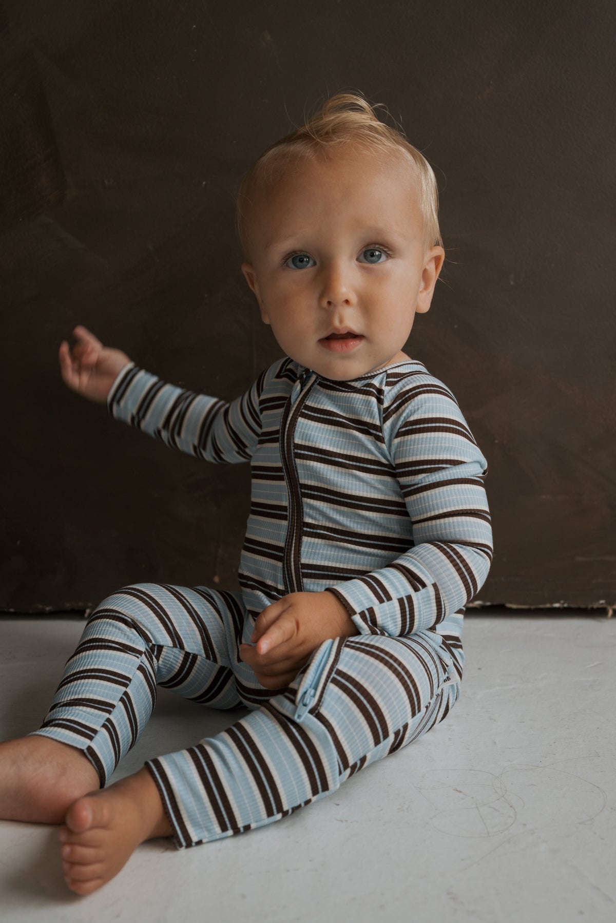 A toddler sits on the floor wearing Bamboo Zip Pajamas | Dean in blue and brown stripes with a front zipper.