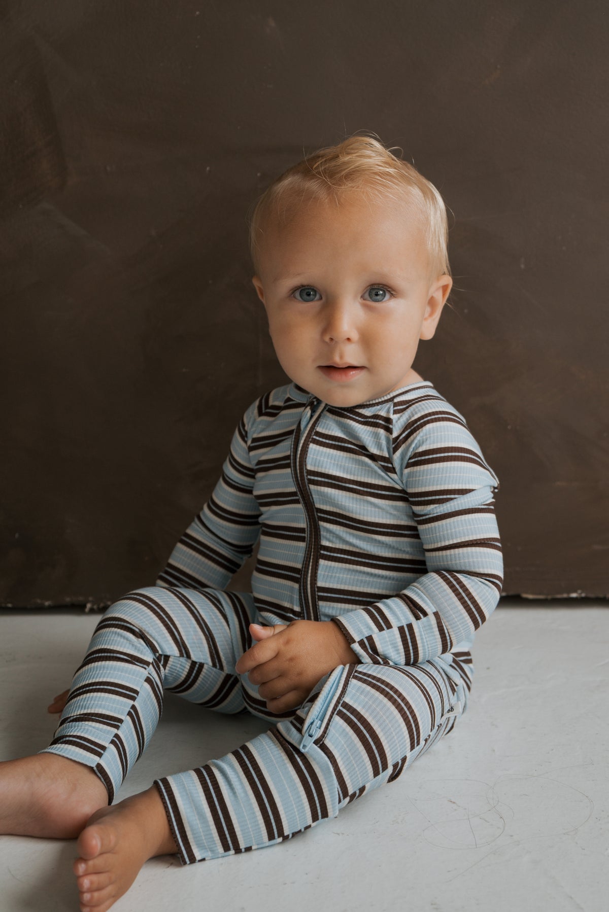 A toddler sits on the floor wearing Dean Bamboo Zip Pajamas by forever french baby, featuring blue and brown striped ribbed bamboo fabric and a front zipper.