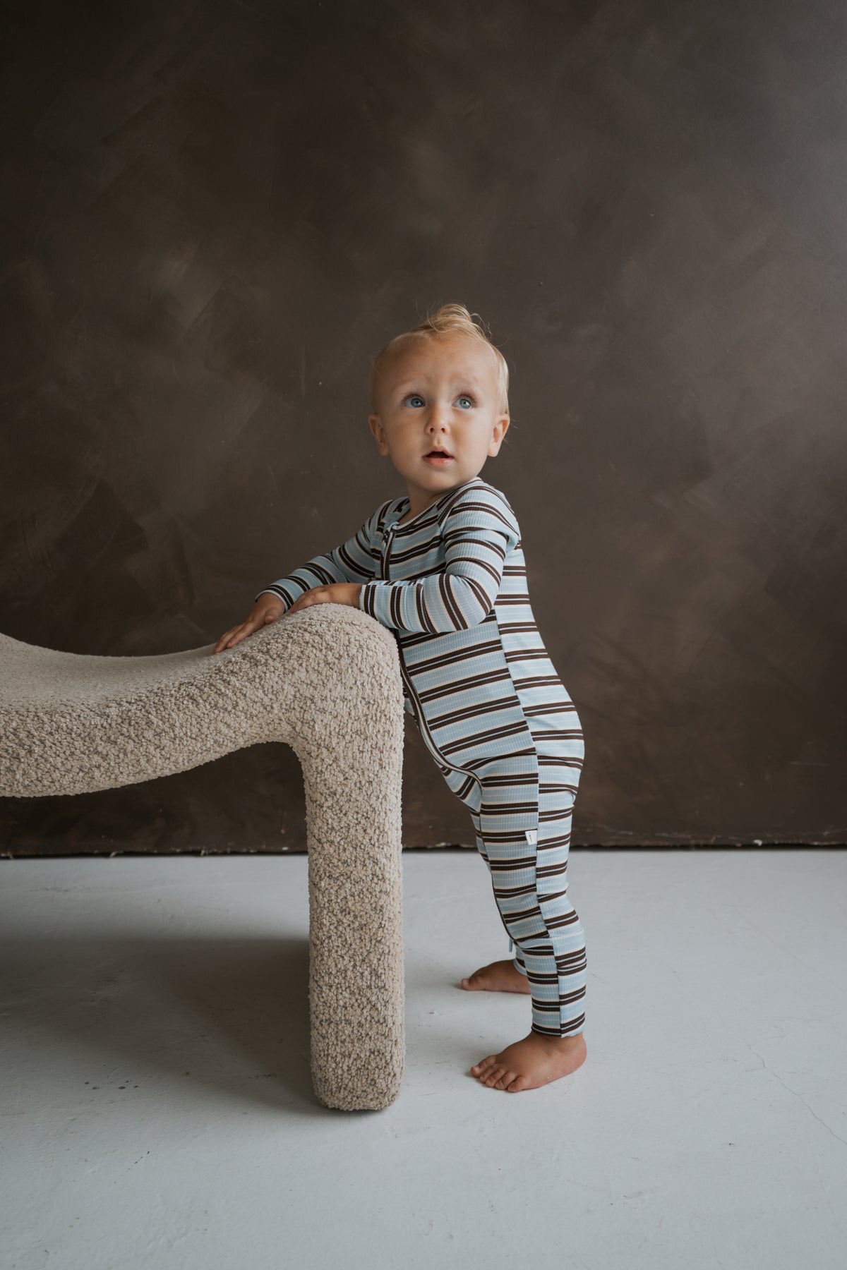 A toddler stands while holding onto a textured bench, wearing blue and brown striped Bamboo Zip Pajamas | Dean by forever french baby.