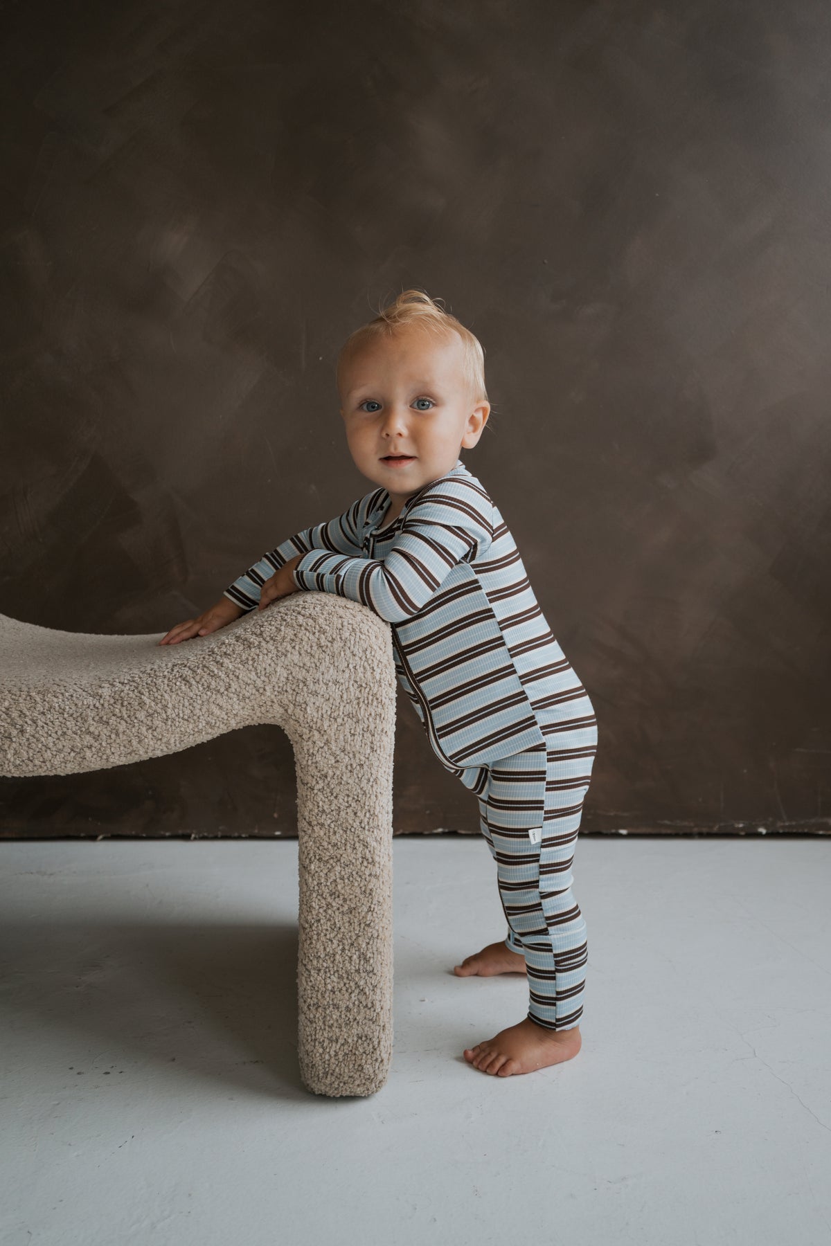 A toddler wearing blue and brown striped ribbed Bamboo Zip Pajamas | Dean stands while leaning against a textured stool.